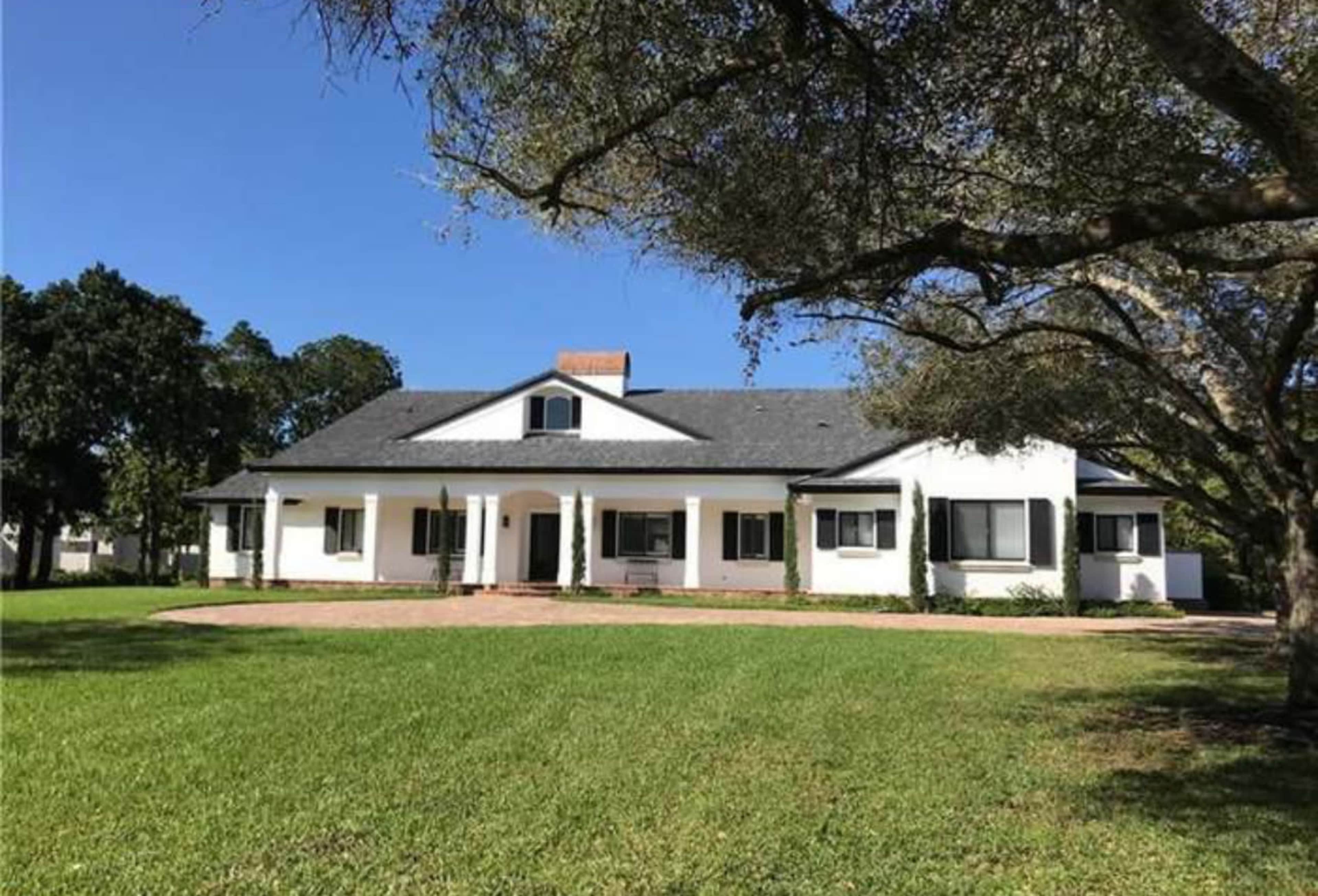 A large white house with a gray roof and a front porch sits on a green lawn, surrounded by trees.