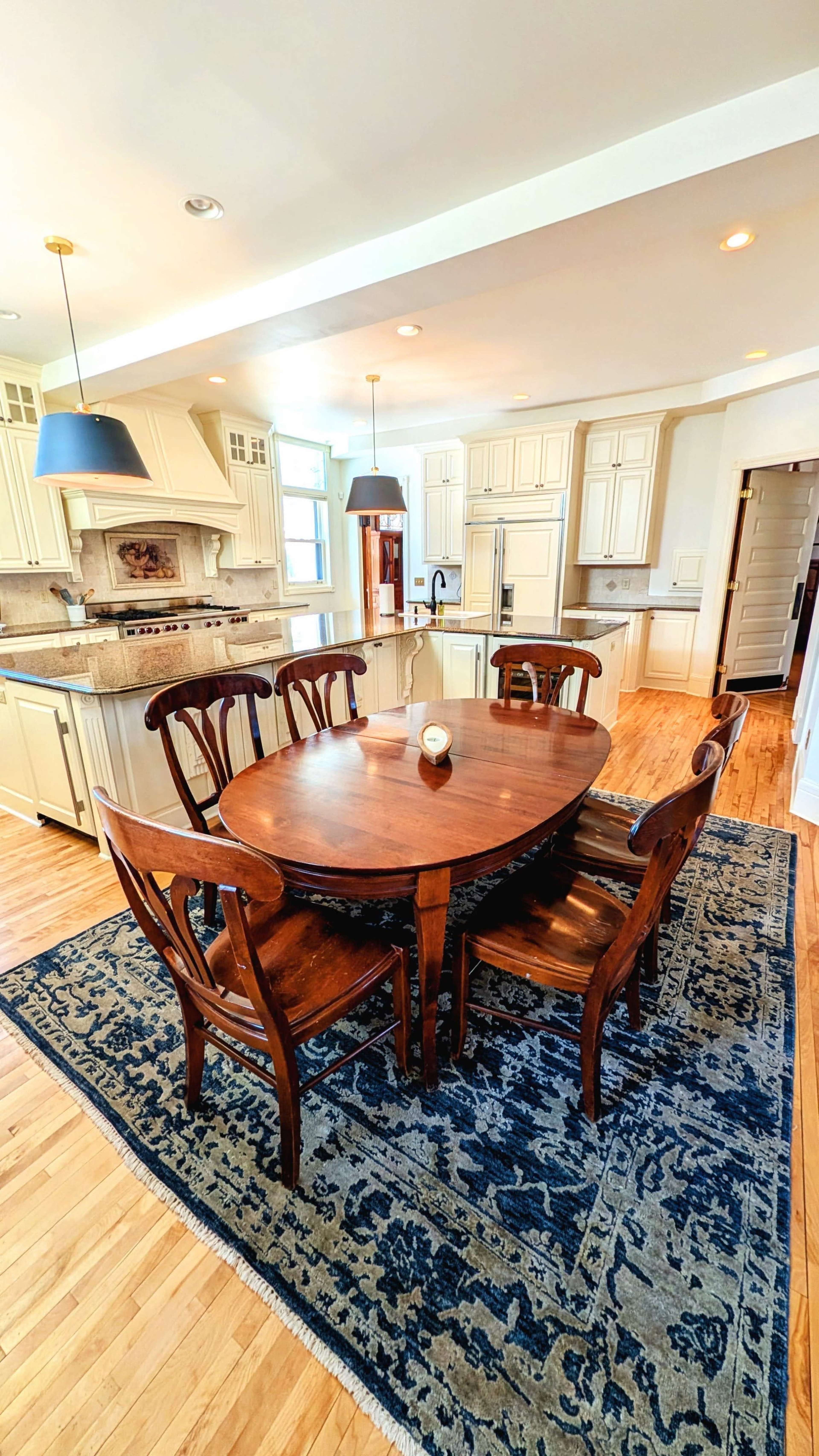 The image shows a spacious kitchen with a round wooden dining table surrounded by six chairs, placed on a decorative rug.