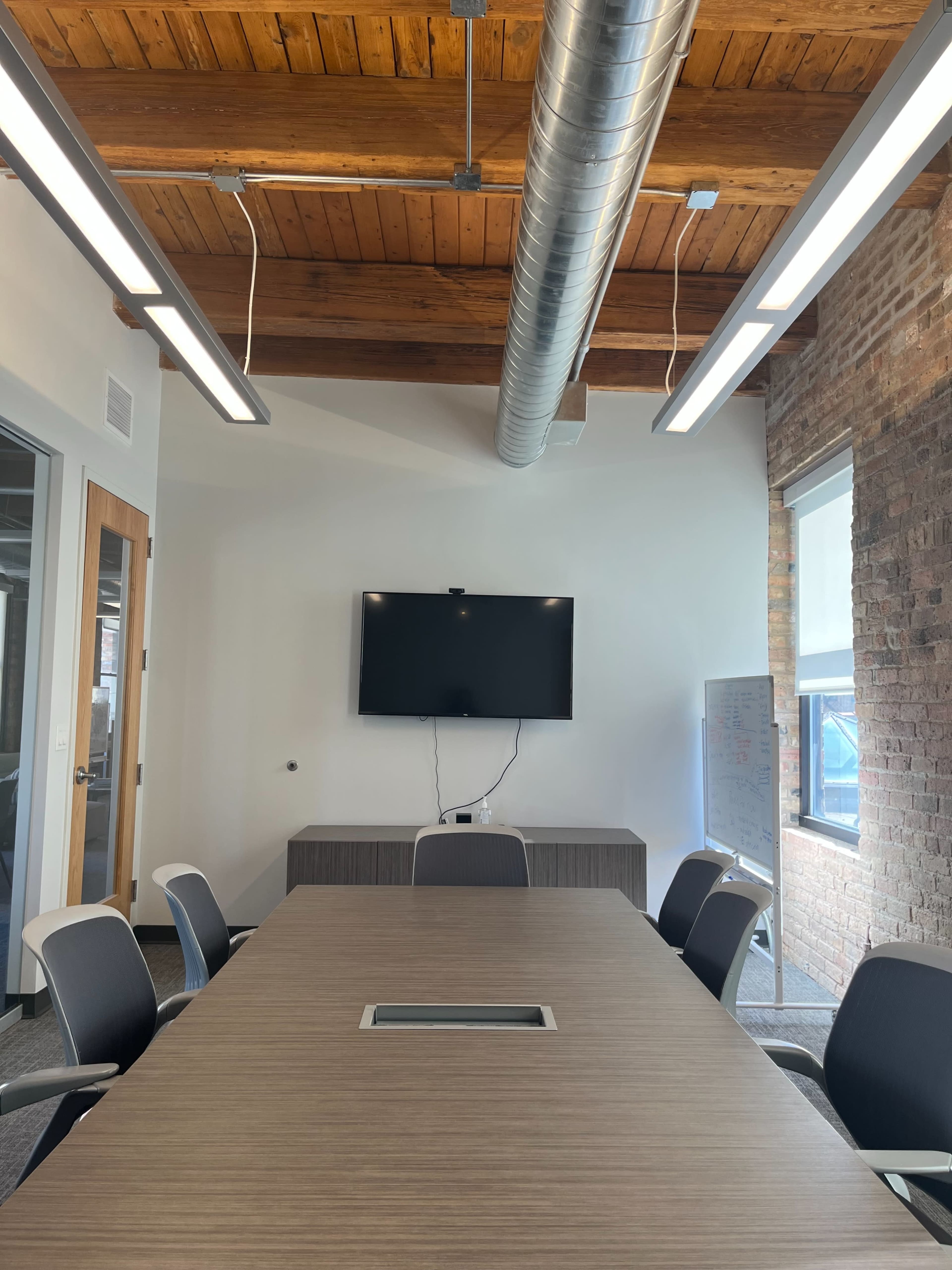 A modern conference room features a large rectangular table, six chairs, a wall-mounted television, and exposed wooden beams in the ceiling.