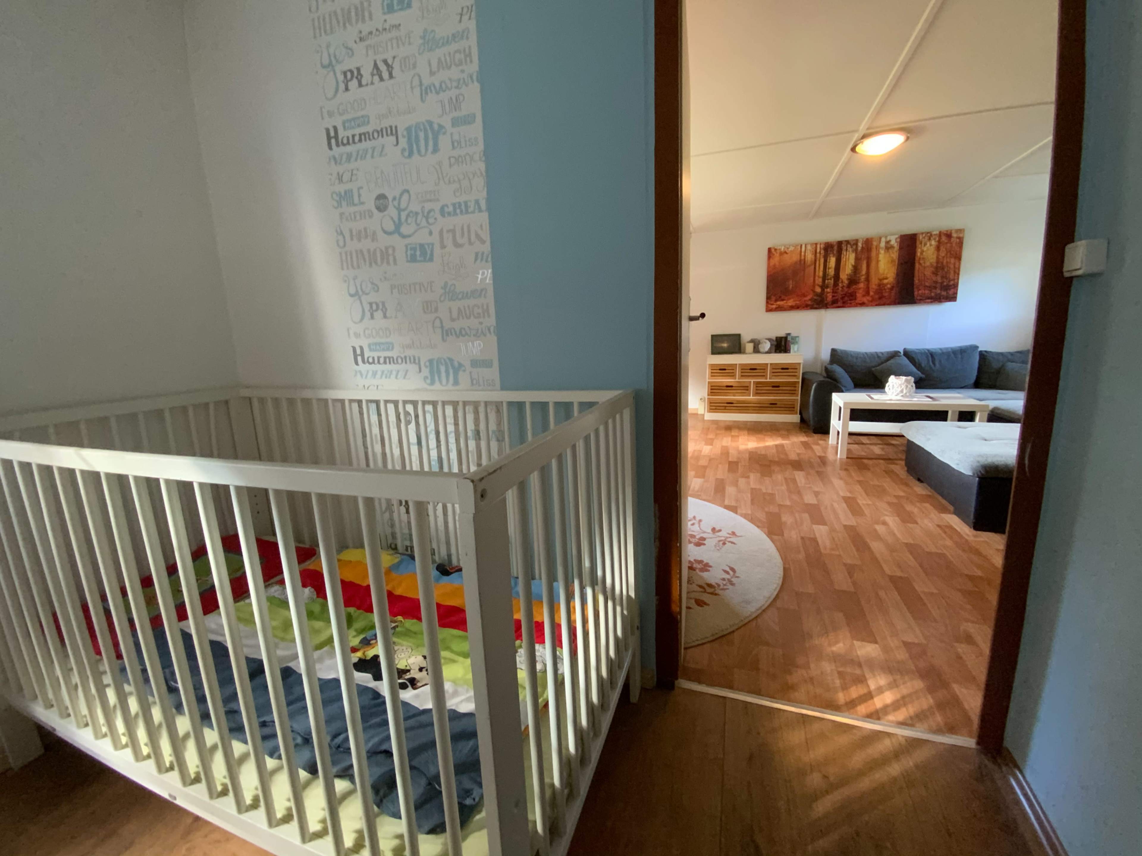 The image shows a small room with a crib on the left and an adjacent living area visible through an open doorway.