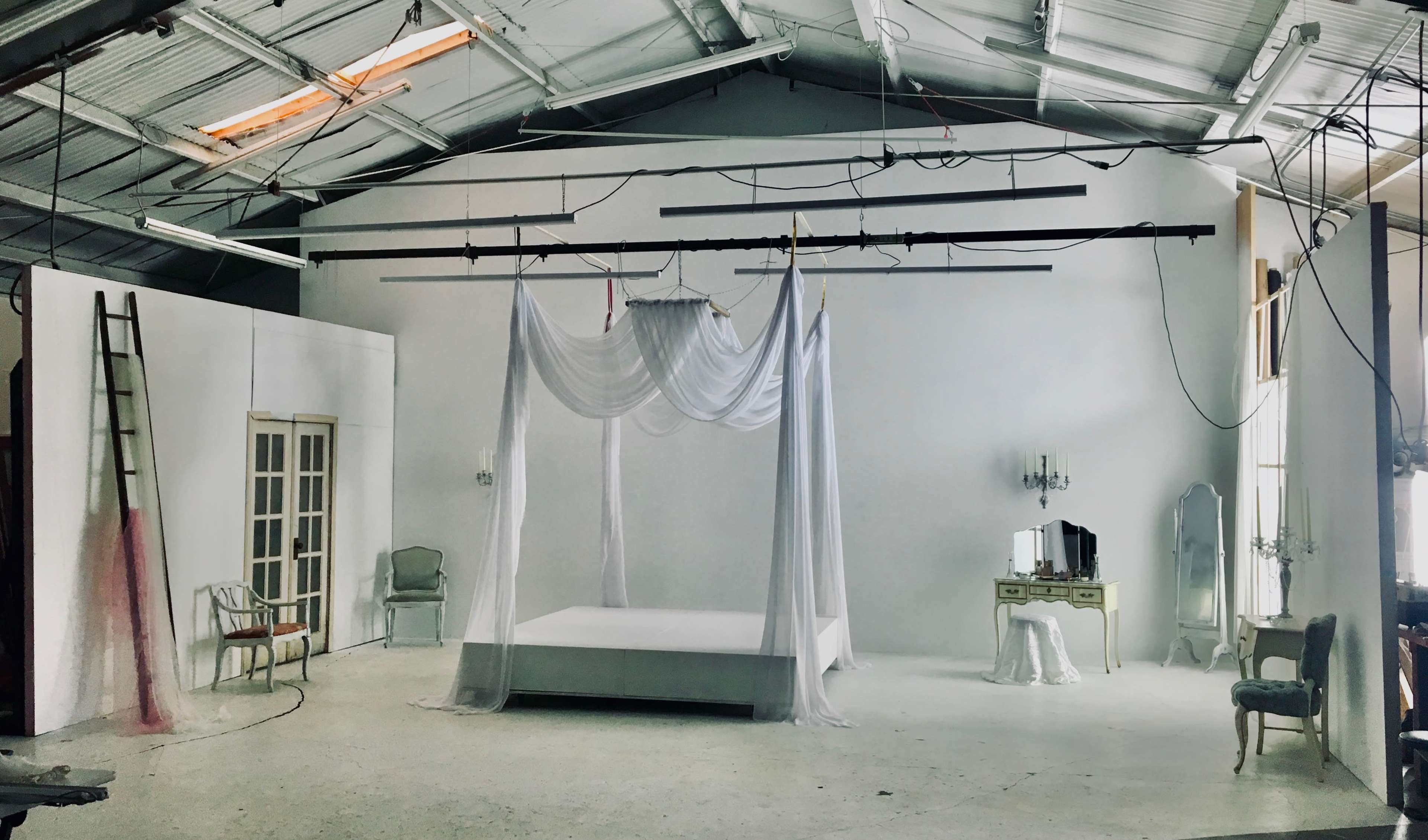 A spacious, empty studio features a white bed draped with sheer fabric, accompanied by vintage furniture and soft lighting.