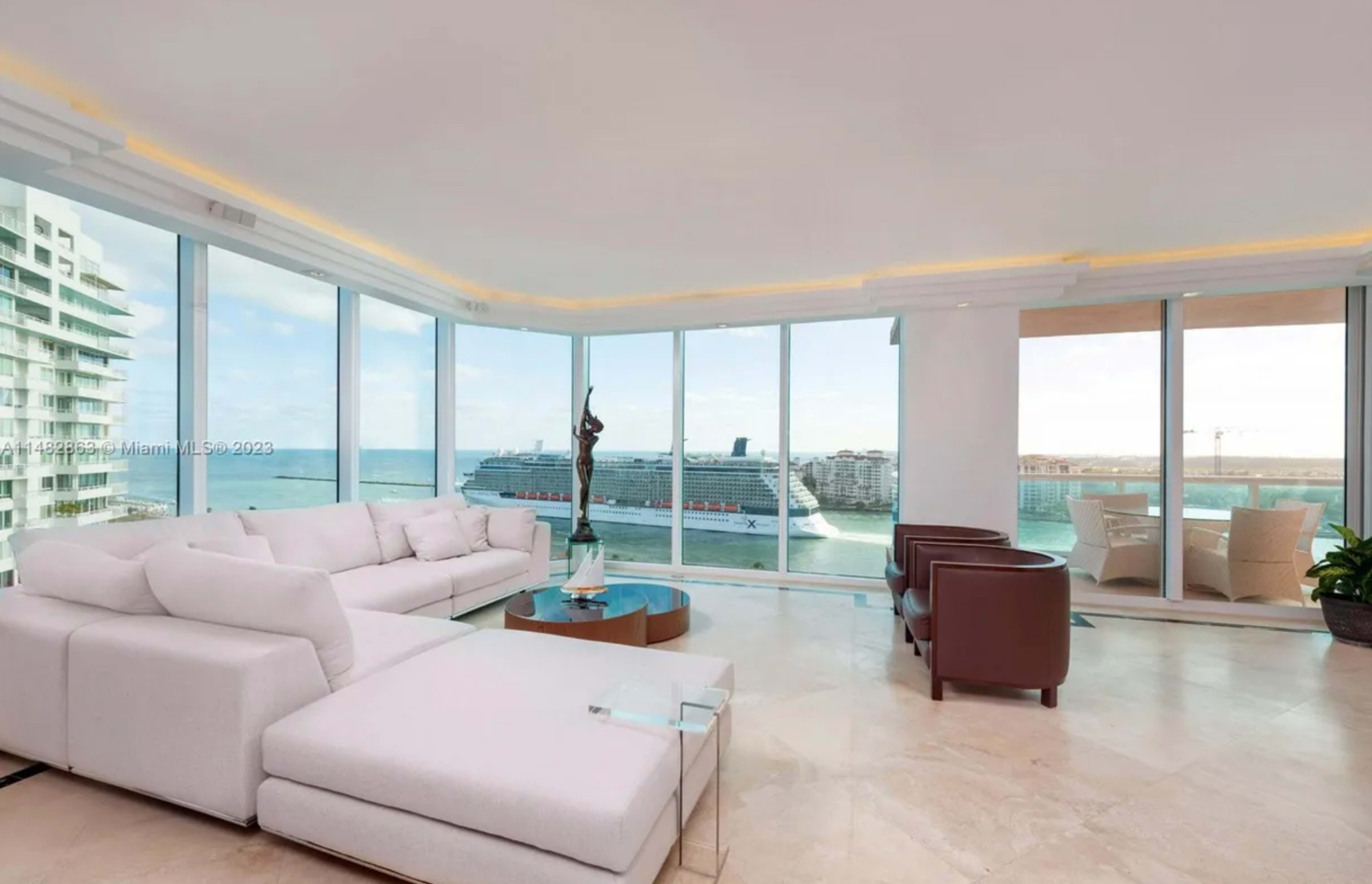 A modern living room features a large white sectional sofa and stunning views of a harbor with a cruise ship through floor-to-ceiling windows.