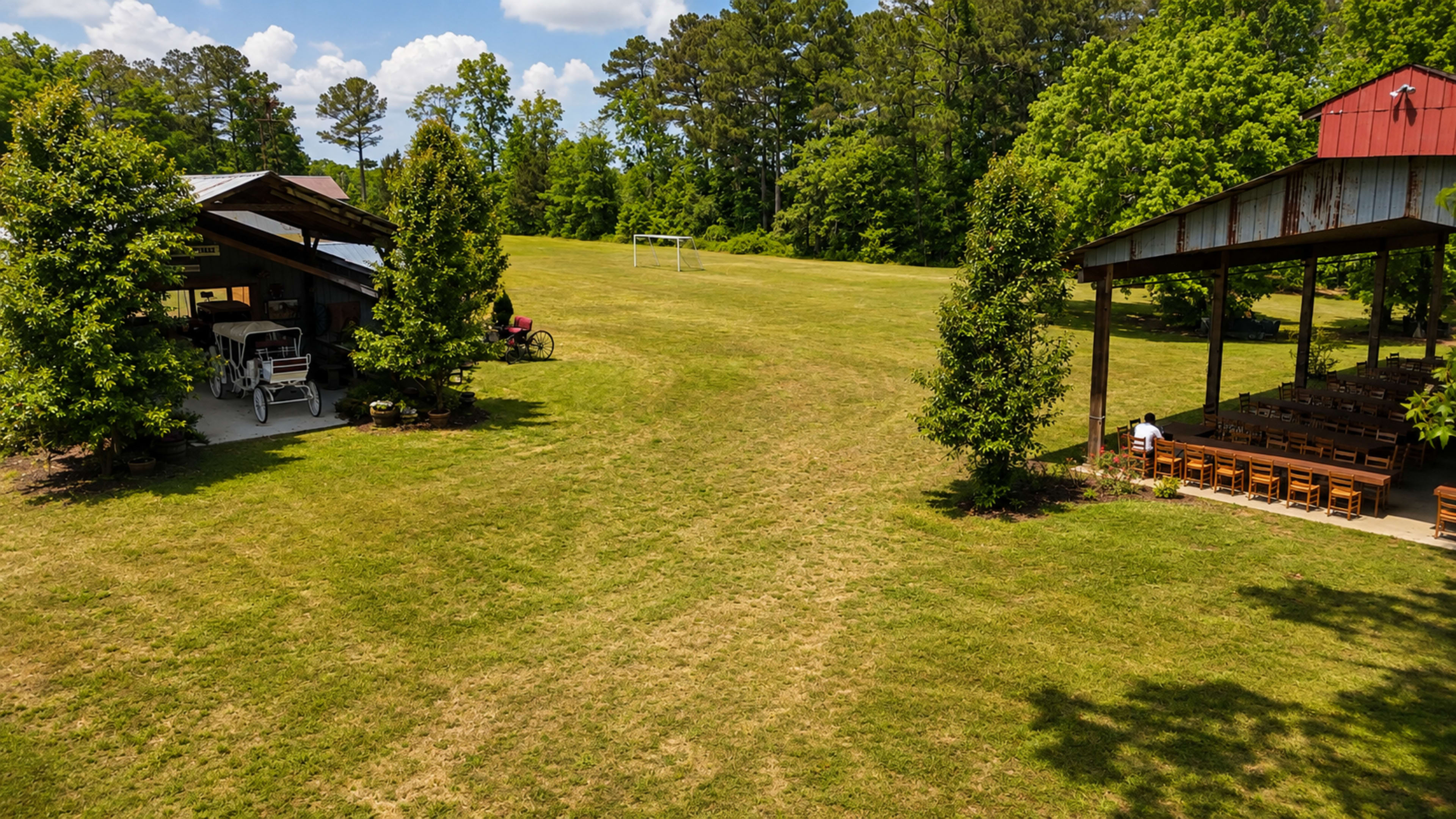 The image shows a spacious green field with a shed on the left and a covered pavilion on the right, surrounded by trees under a partly cloudy sky.