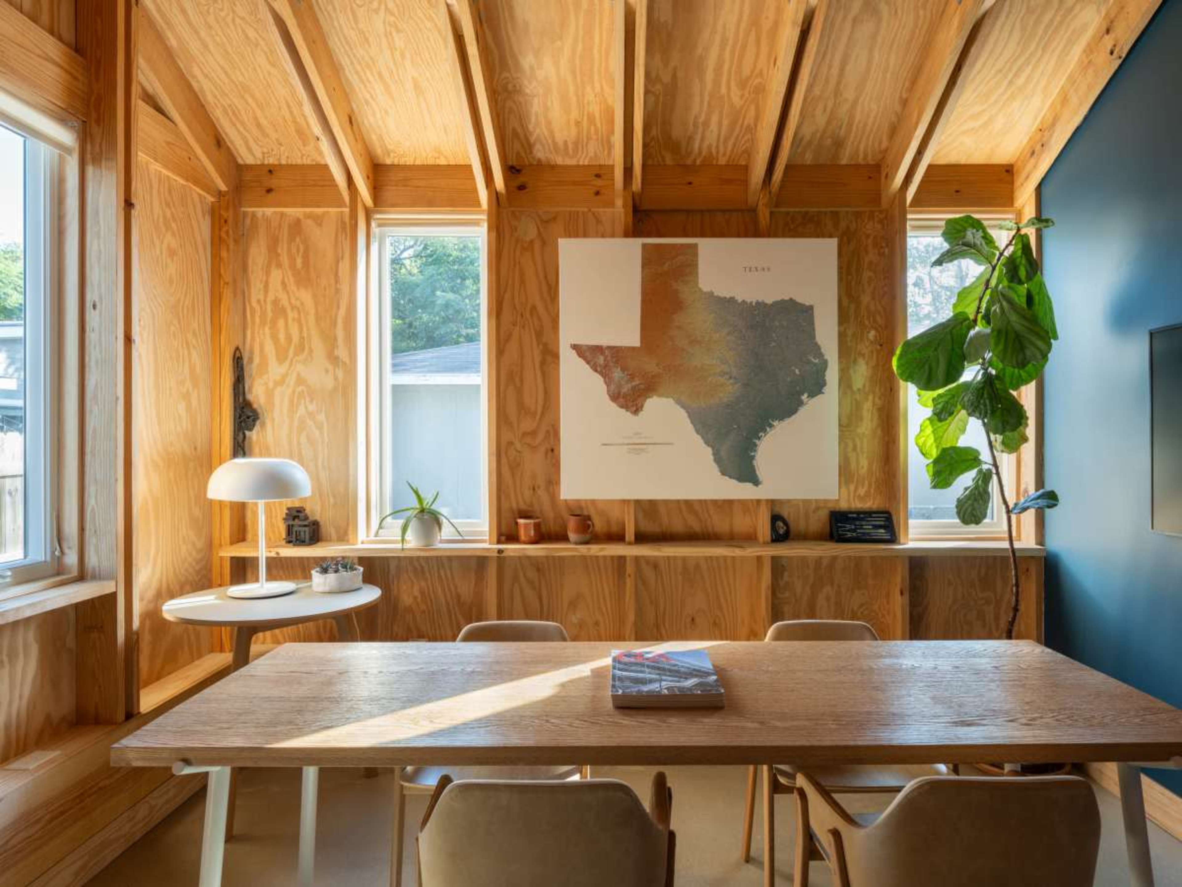 The image shows a wooden room with a large map of Texas on the wall, a dining table, chairs, a modern lamp, and plants near the windows.
