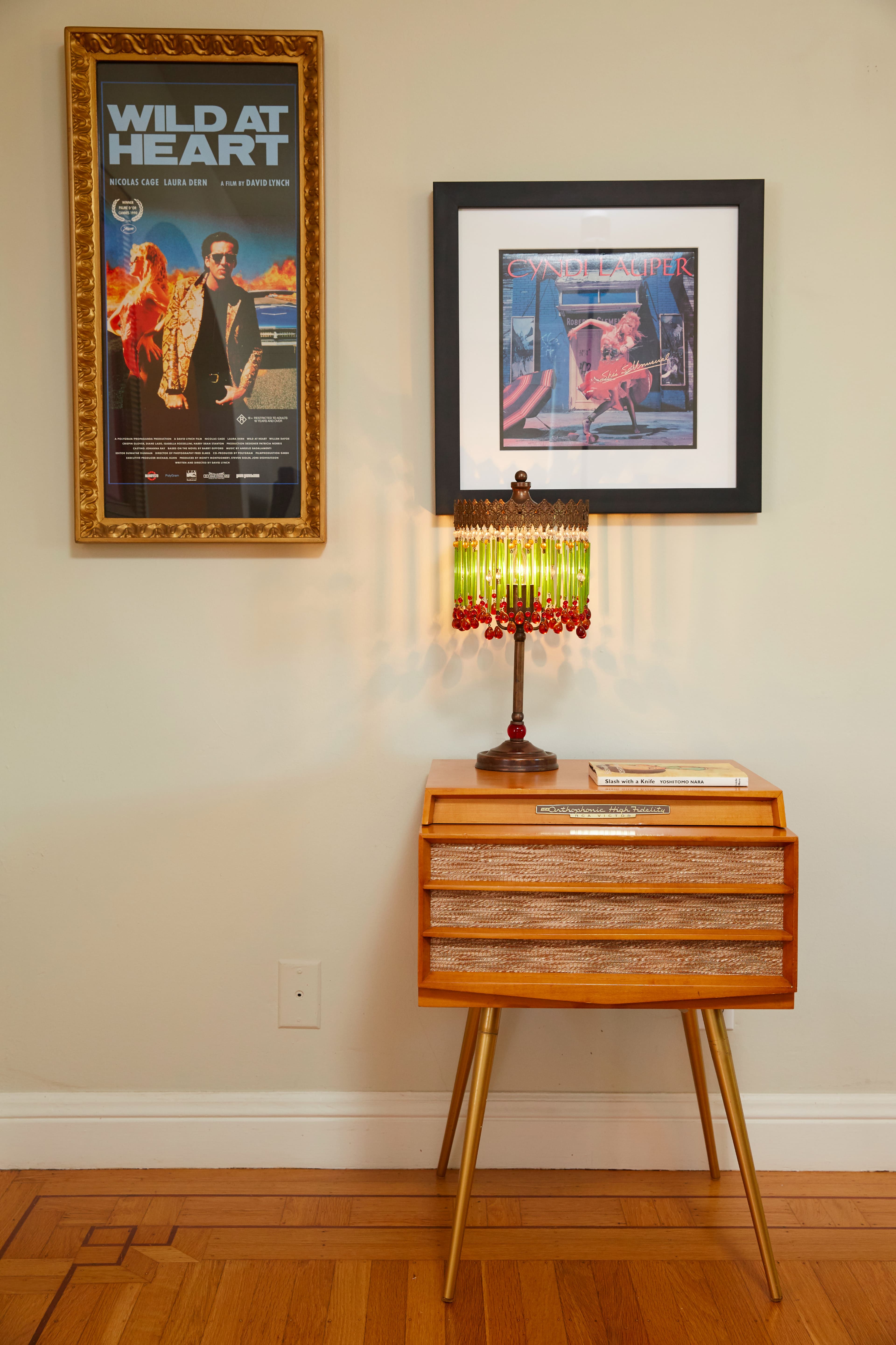 A wooden side table with a patterned lamp, a framed poster of "Wild at Heart" on the wall, and a framed artwork hanging above.