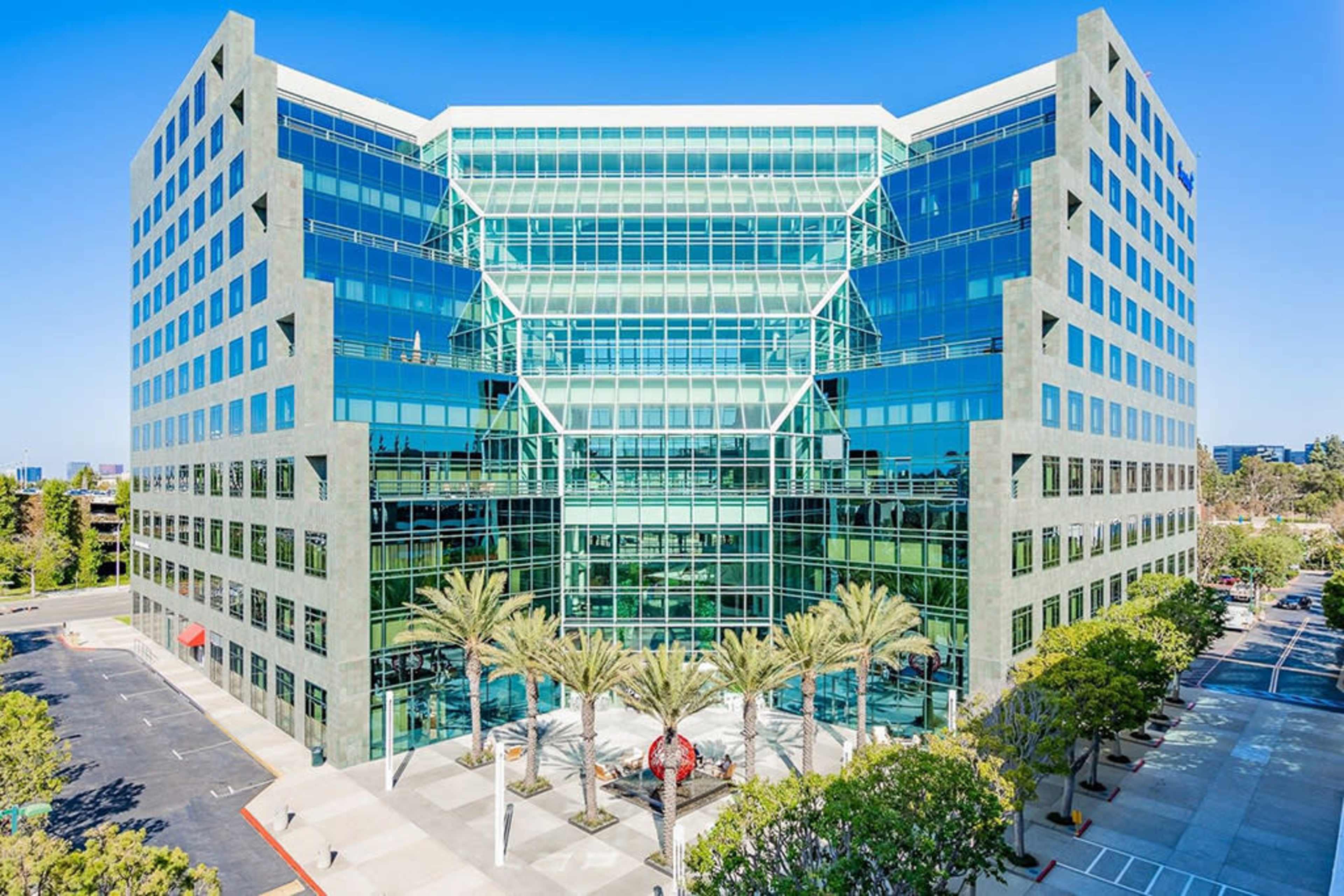 The image shows a modern, multi-story office building with a glass facade surrounded by palm trees and a parking area.