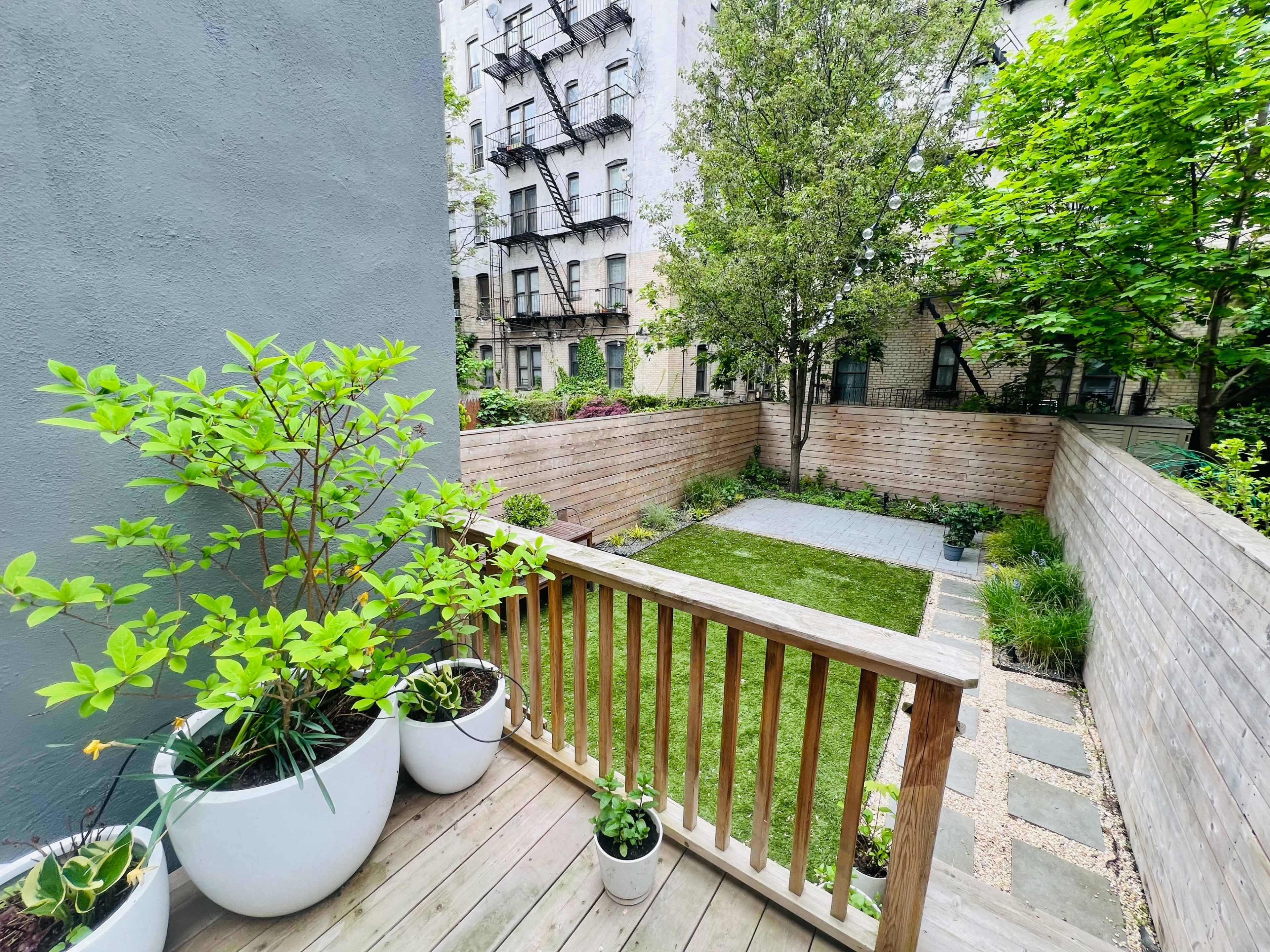 A wooden deck overlooking a small garden with a grassy area and neatly arranged plants against a backdrop of buildings.