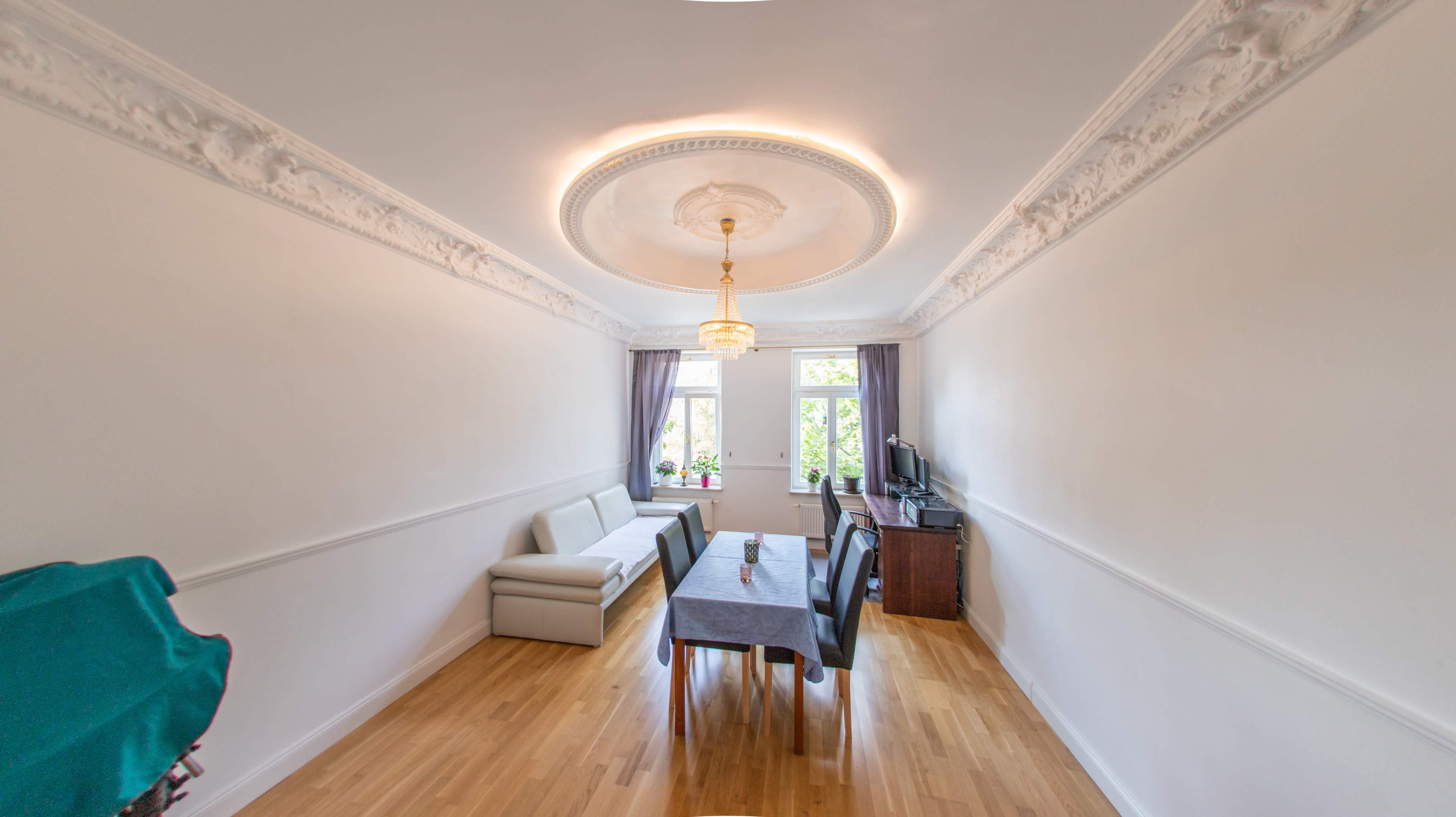 The image depicts a bright room featuring a table with chairs, a sofa against one wall, a television unit, and large windows with drapes.