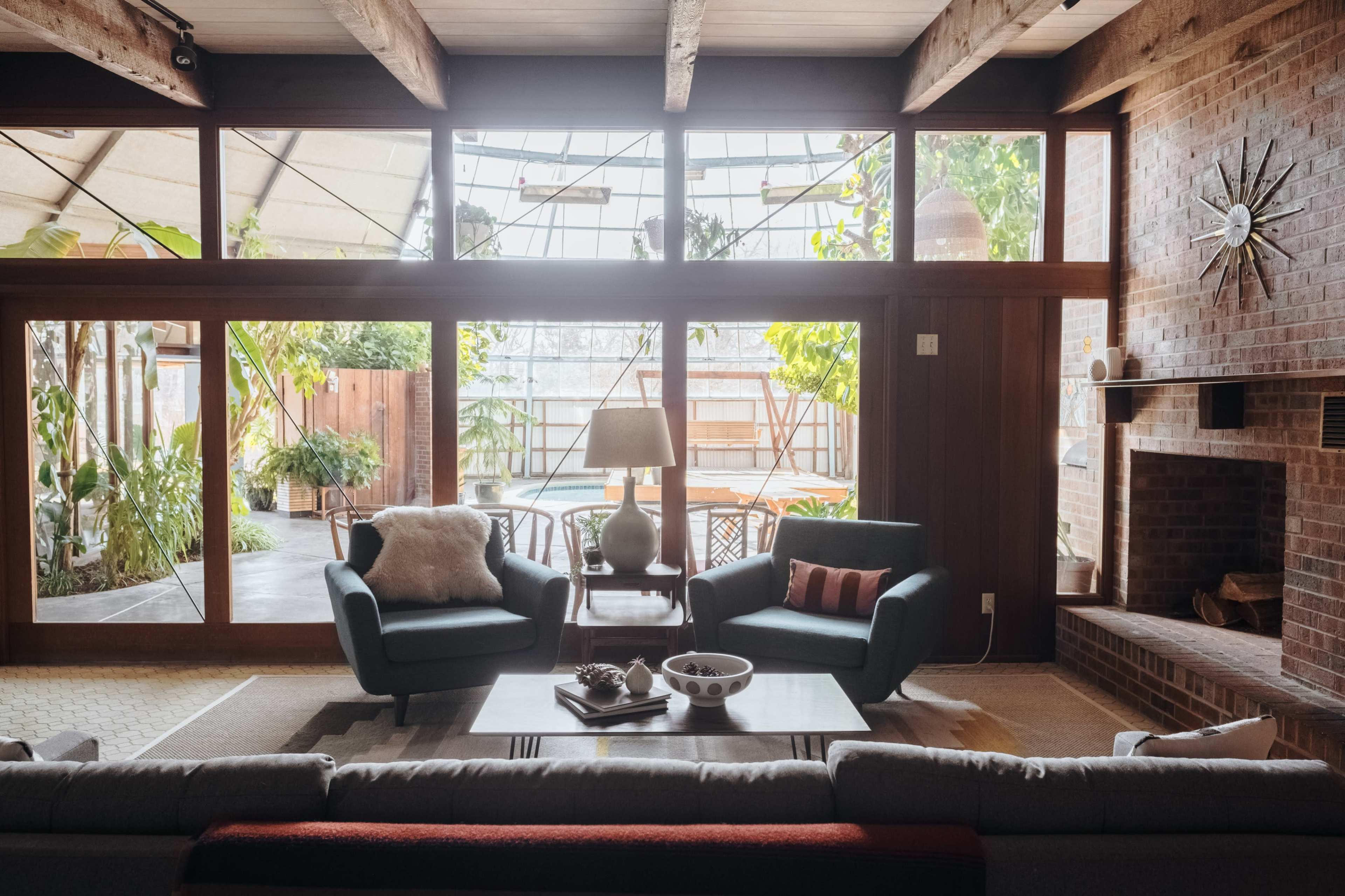 The living room features two teal armchairs facing a coffee table, with large windows providing a view of a plant-filled outdoor space.