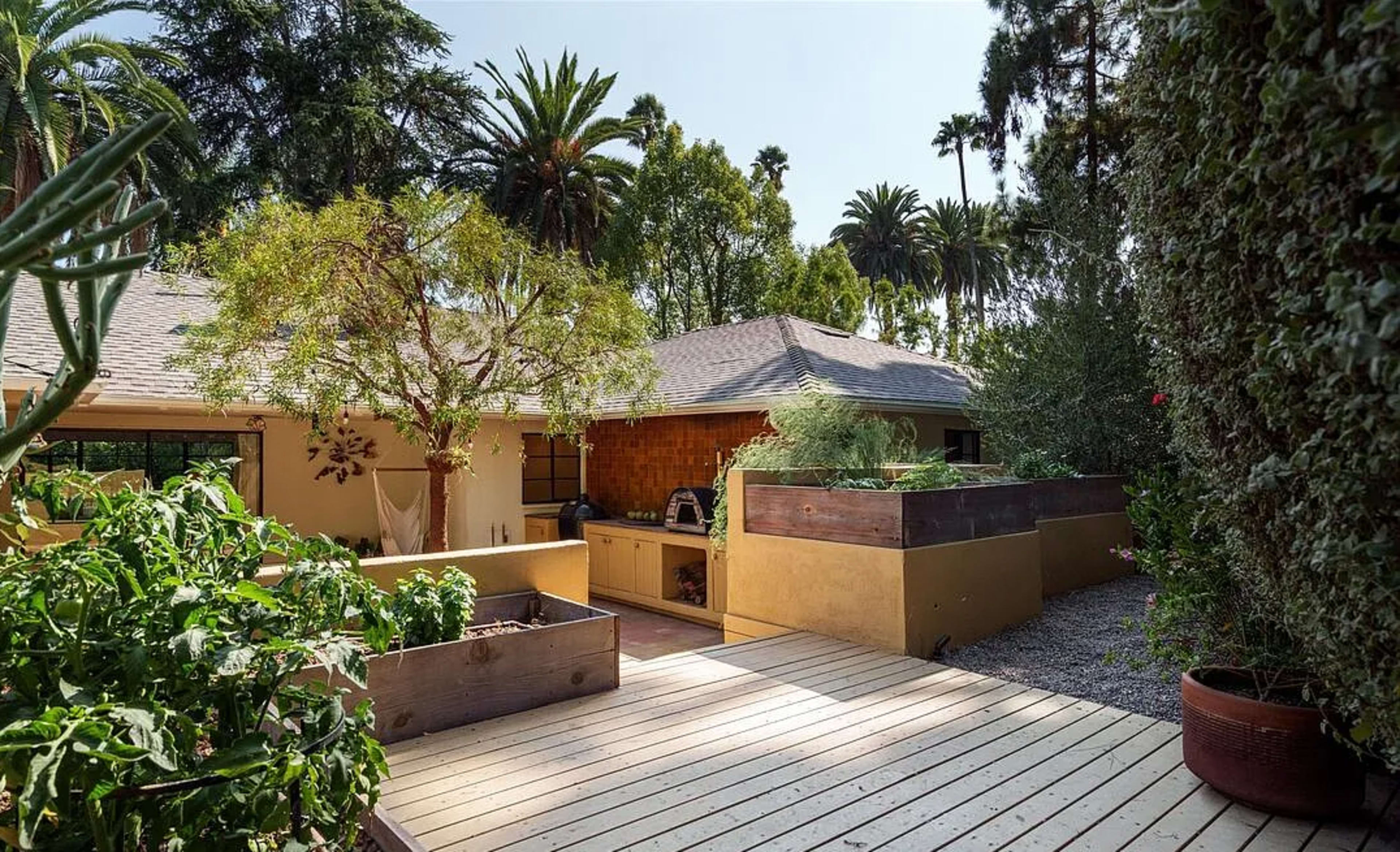 The image shows a landscaped backyard with wooden decking, garden beds, and a house surrounded by palm trees.