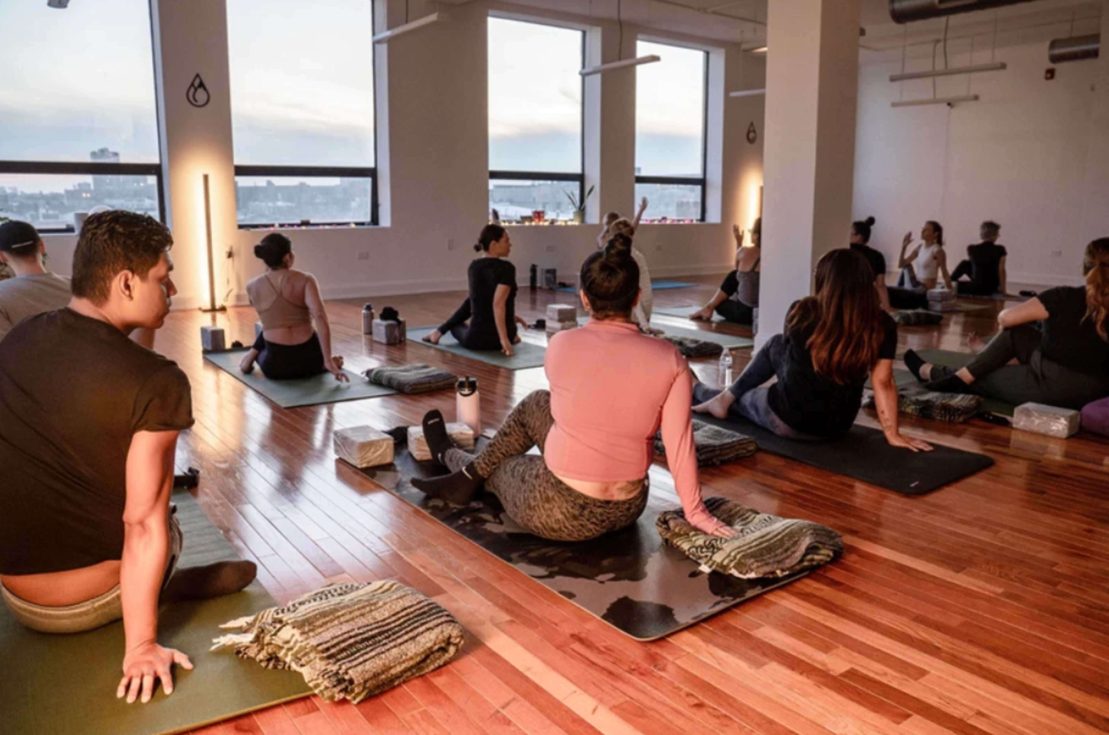 A group of people participates in a yoga class in a spacious studio with large windows and wooden flooring.