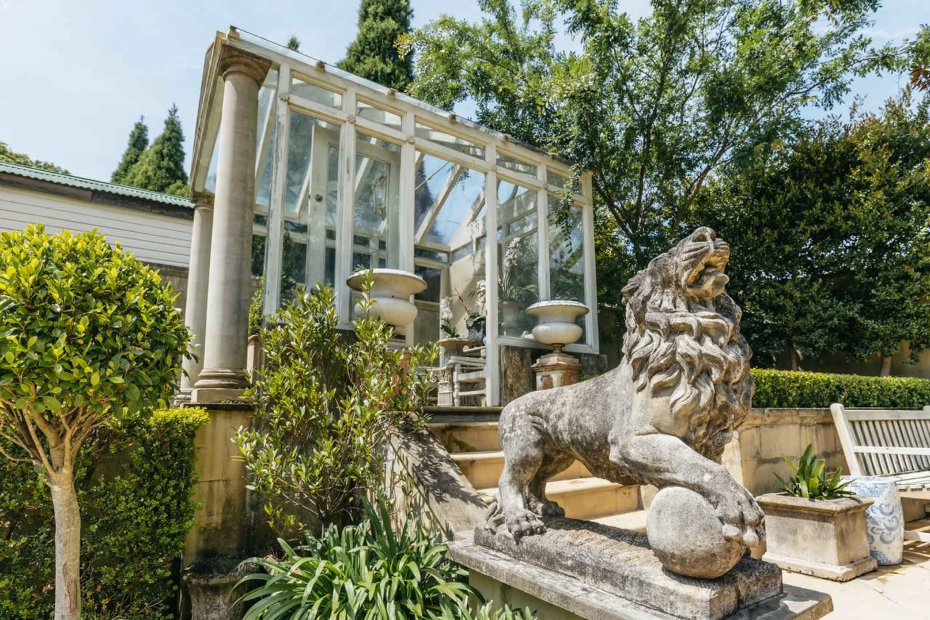 A lion statue rests on a stone pedestal in front of a glass greenhouse surrounded by neatly trimmed bushes and trees.