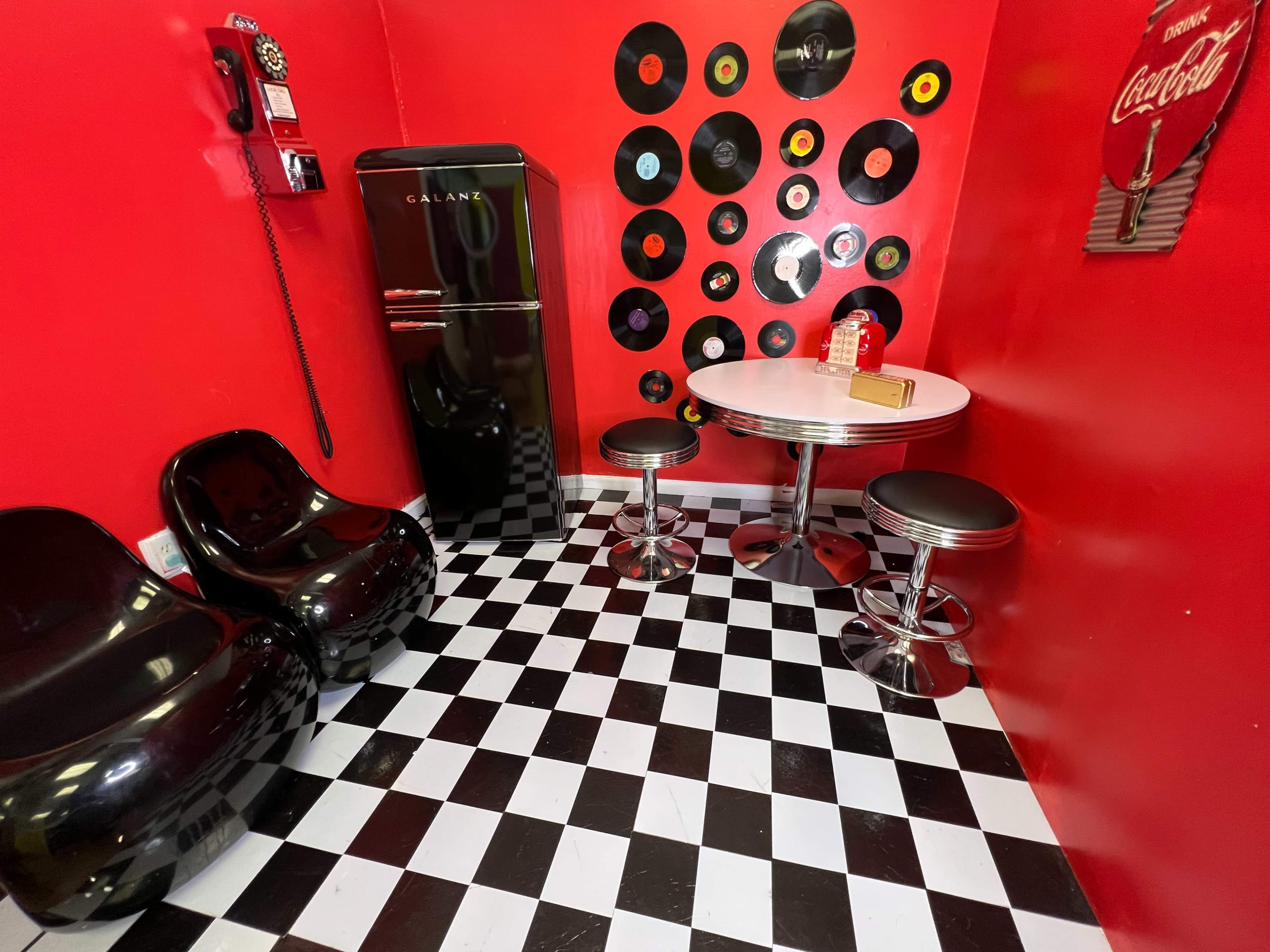 The scene features a small room with red walls adorned with vinyl records, a black refrigerator, two black chairs, and a round table with black and silver stools on a black and white checkered floor.