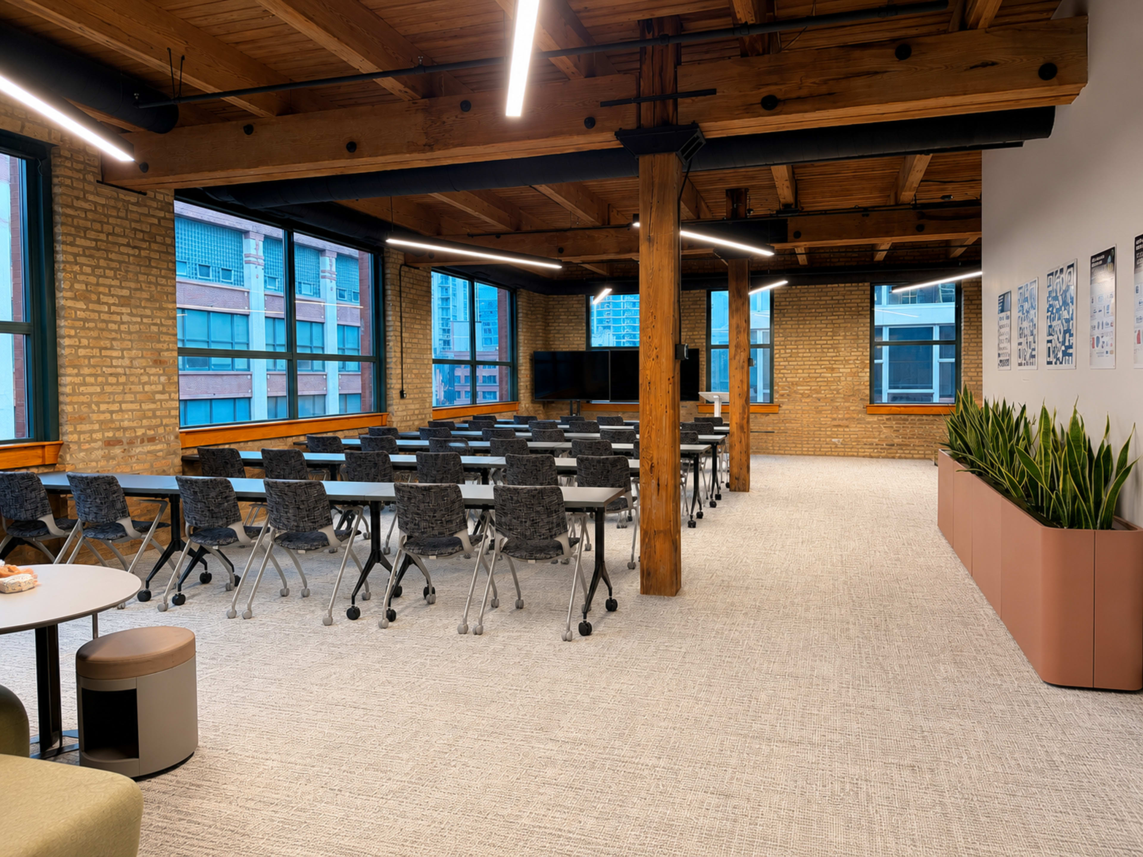 The image shows a modern office space featuring a large meeting area with rows of tables and chairs, exposed wooden beams, and large windows.