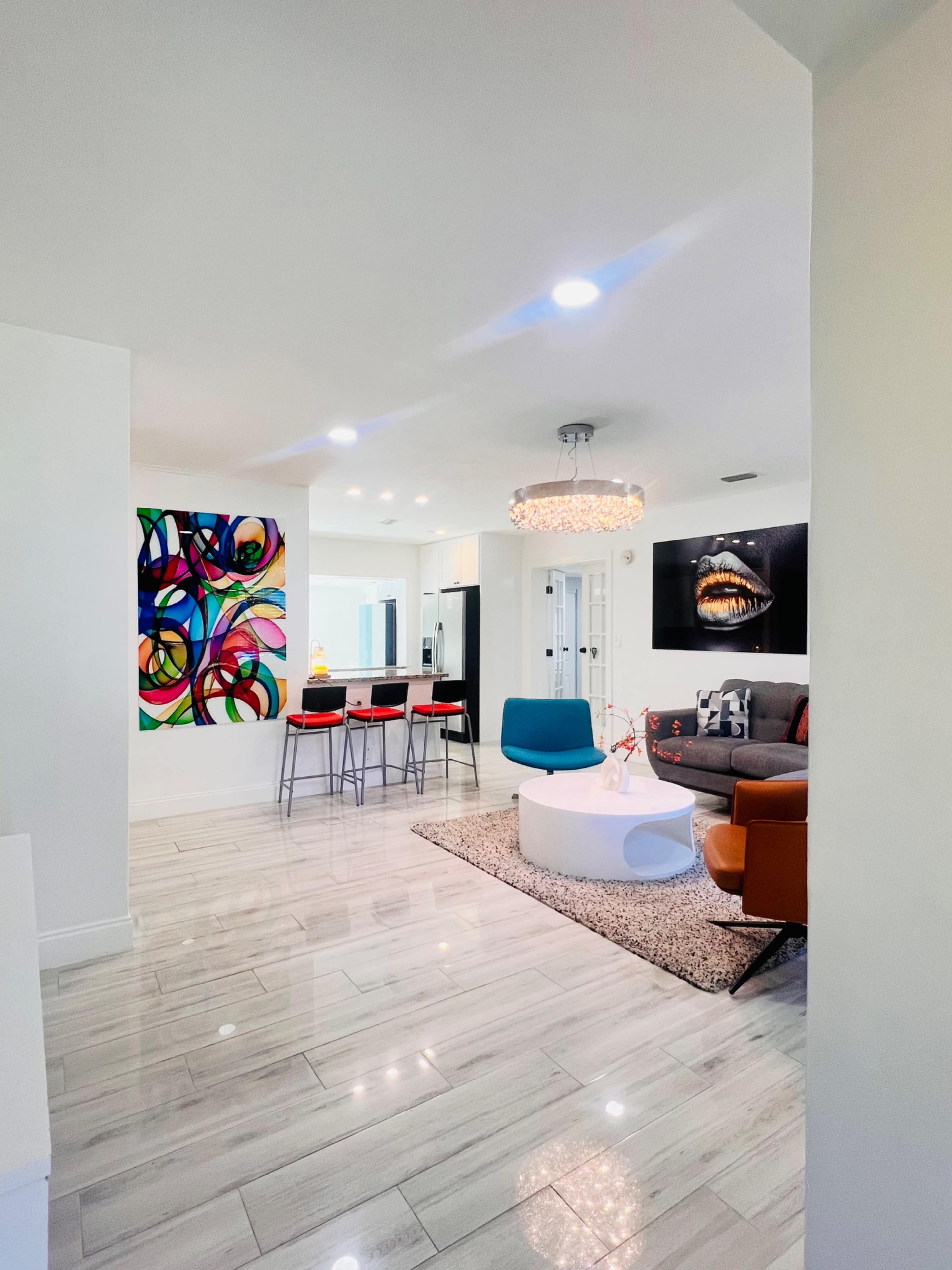 The image shows a modern living area with a bright chandelier, colorful wall art, stylish furniture, and a small dining area with bar seating.