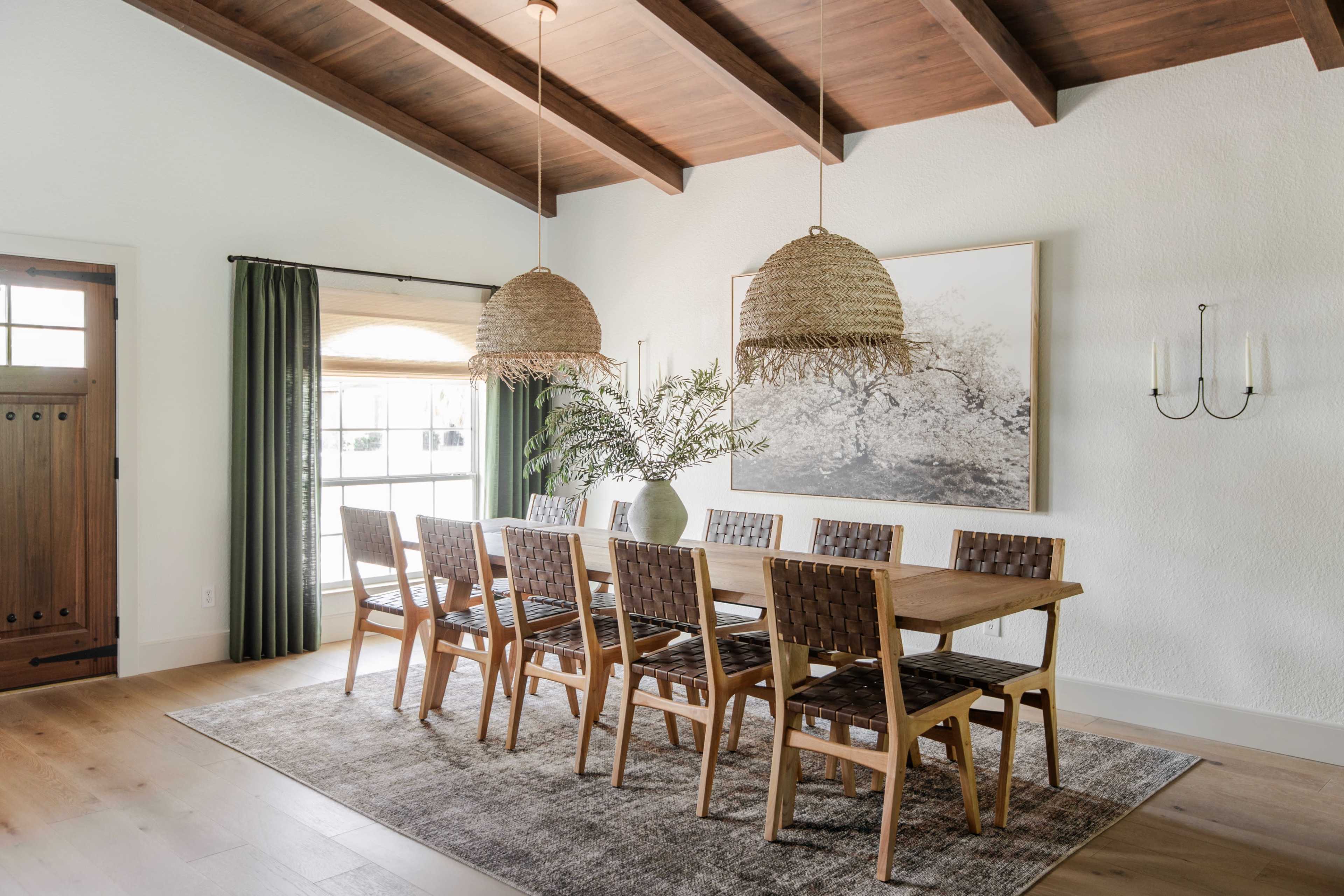 A spacious dining room features a long wooden table surrounded by eight woven chairs, with large pendant lights hanging above and a framed artwork on the wall.
