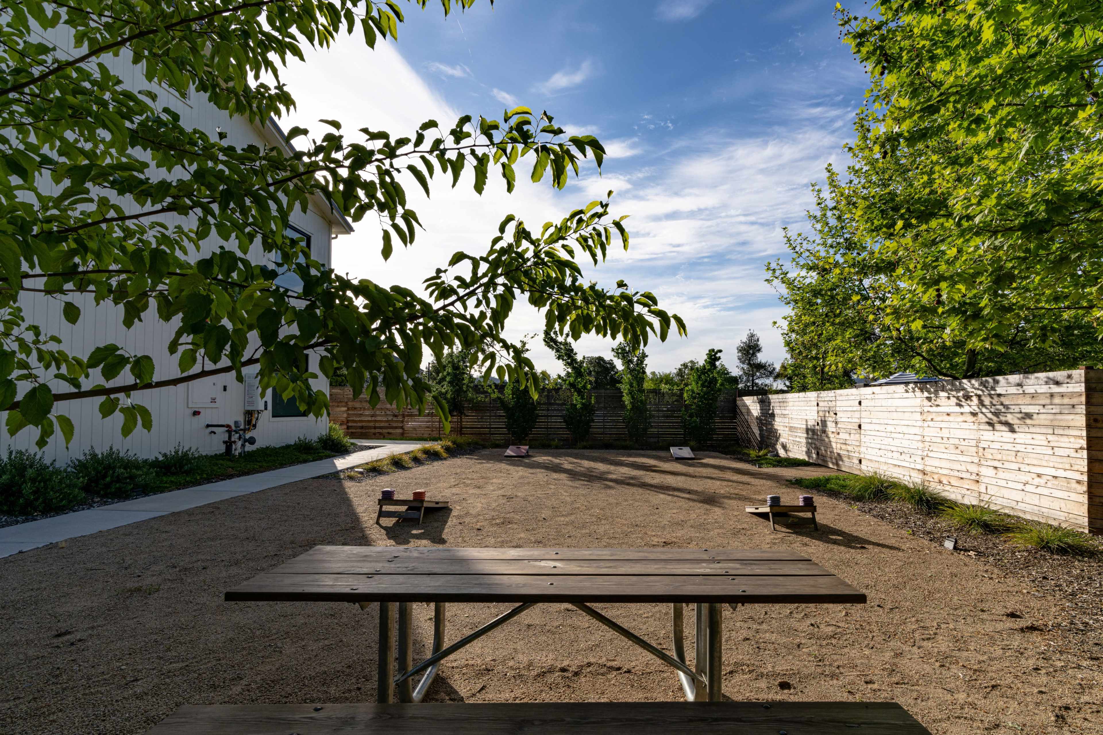 A wooden picnic table is placed in a sandy area surrounded by greenery and a wooden fence on a clear day.