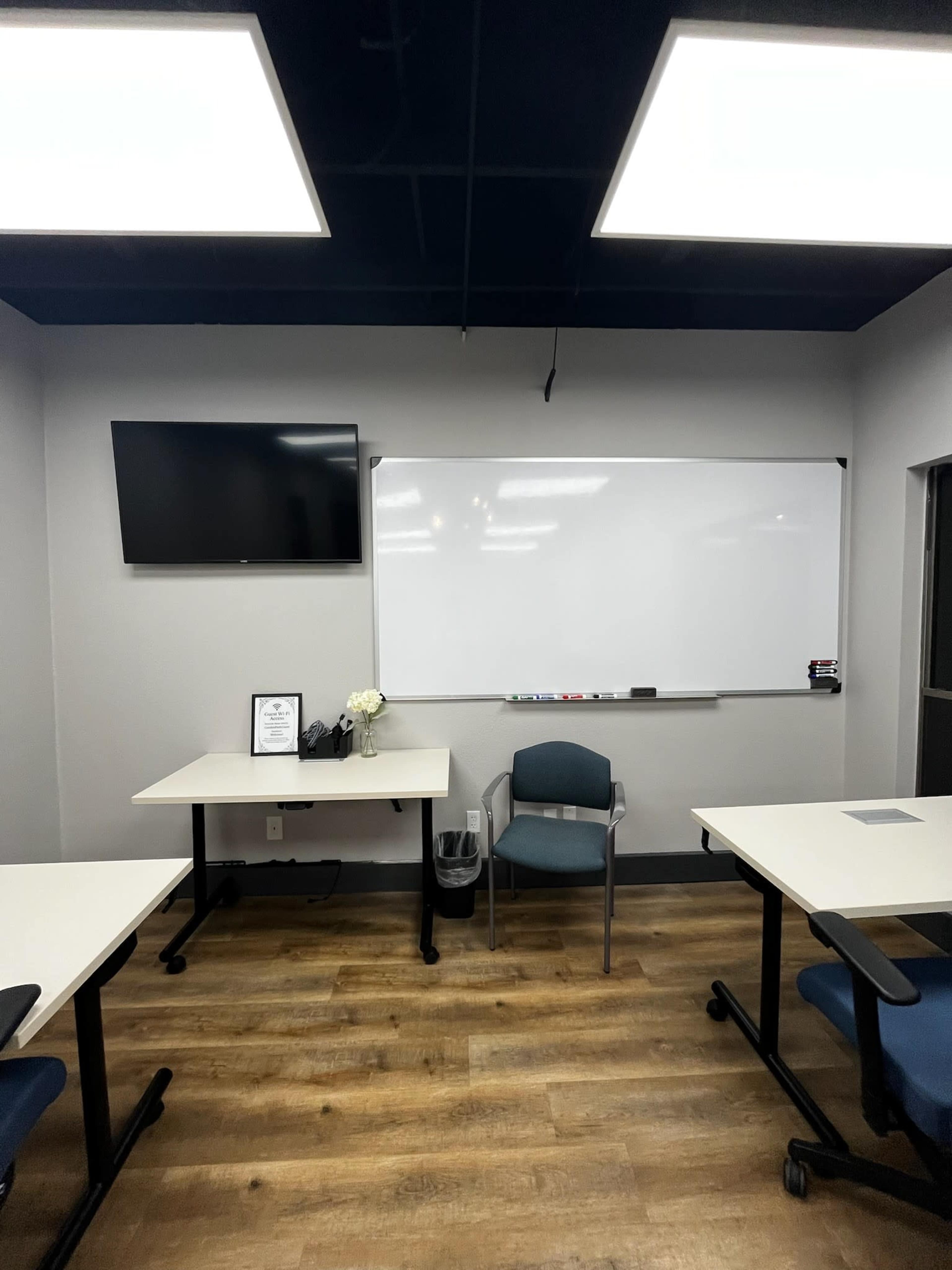 The image shows a meeting room with a television mounted on the wall, a dry-erase board, a single chair, and several tables arranged around a wood laminate floor.