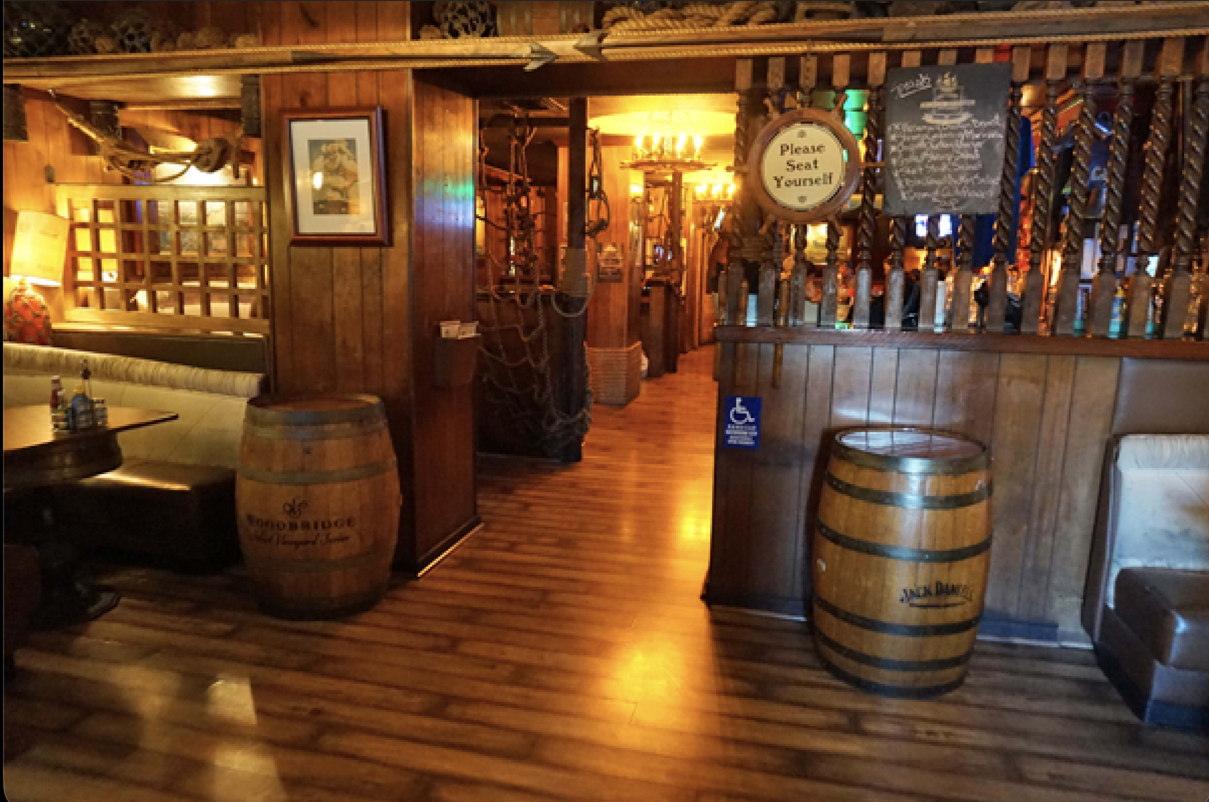 The image shows a rustic wooden interior of a restaurant, featuring barrels and nautical decor with a pathway leading toward a brightly lit area.