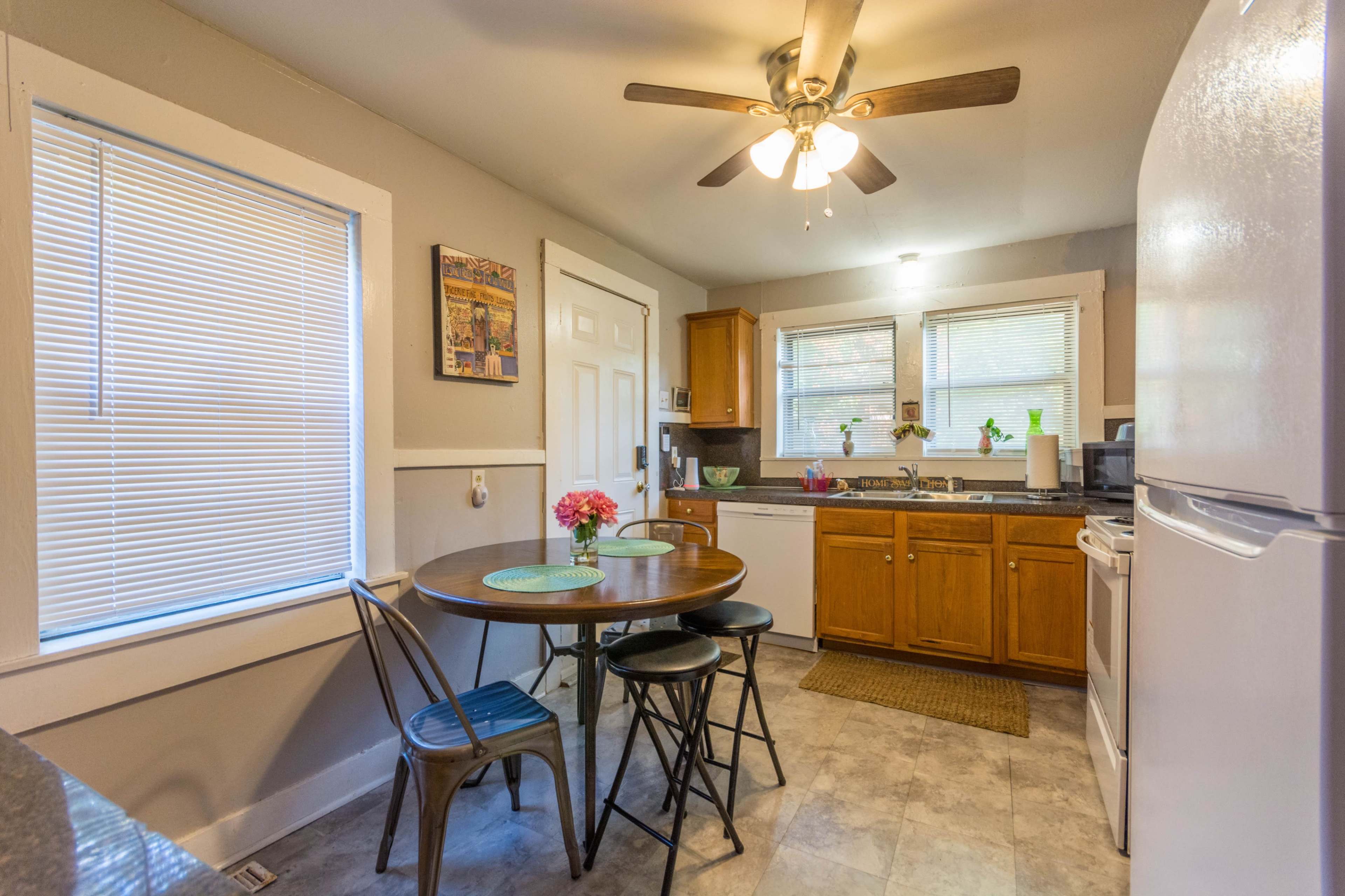 A small kitchen features a round table with two chairs, wooden cabinets, a refrigerator, and a ceiling fan.