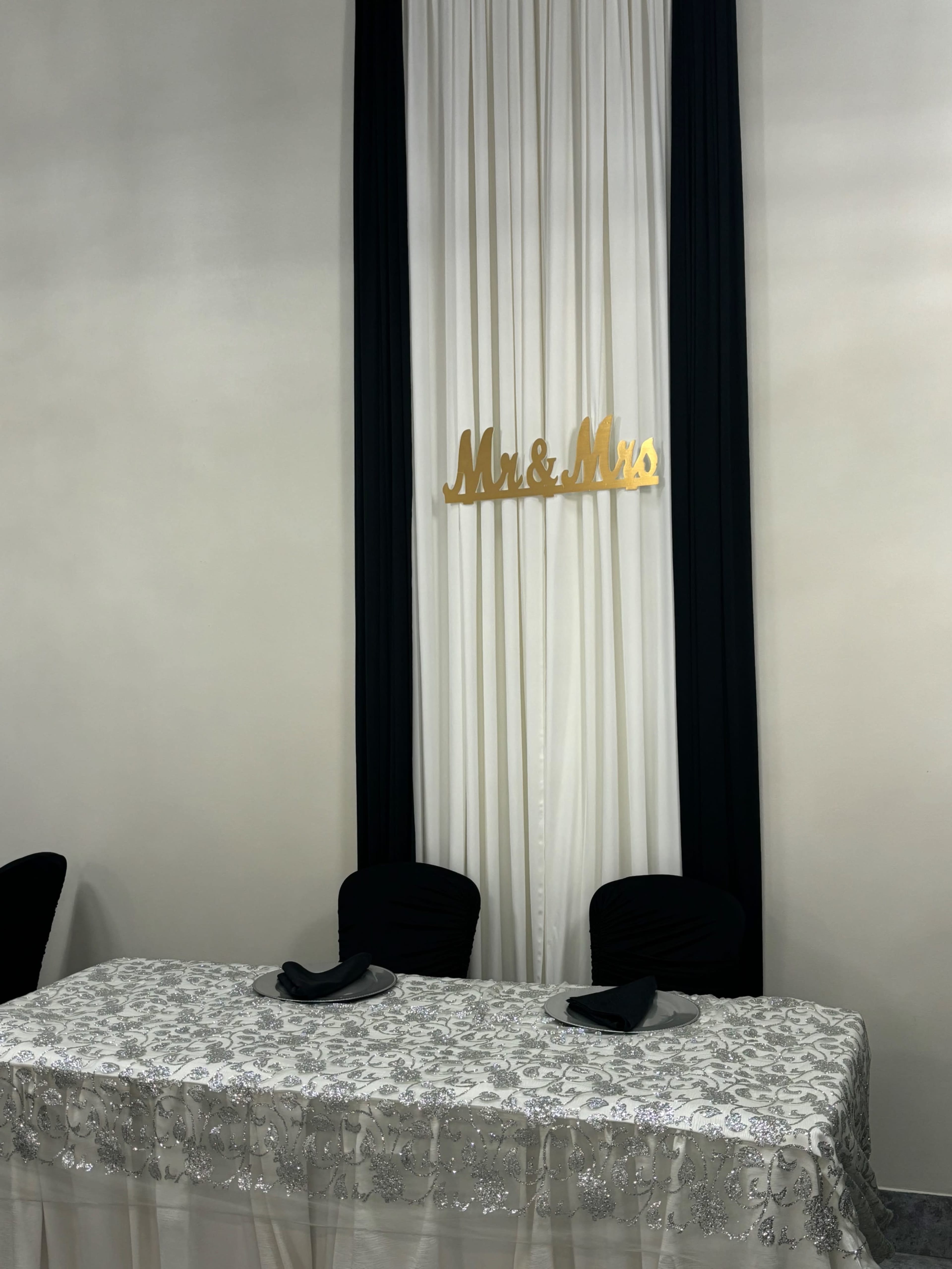 A table is set for a wedding reception, adorned with a silver tablecloth and a "Mr. & Mrs." sign hanging above a backdrop of white and navy drapery.