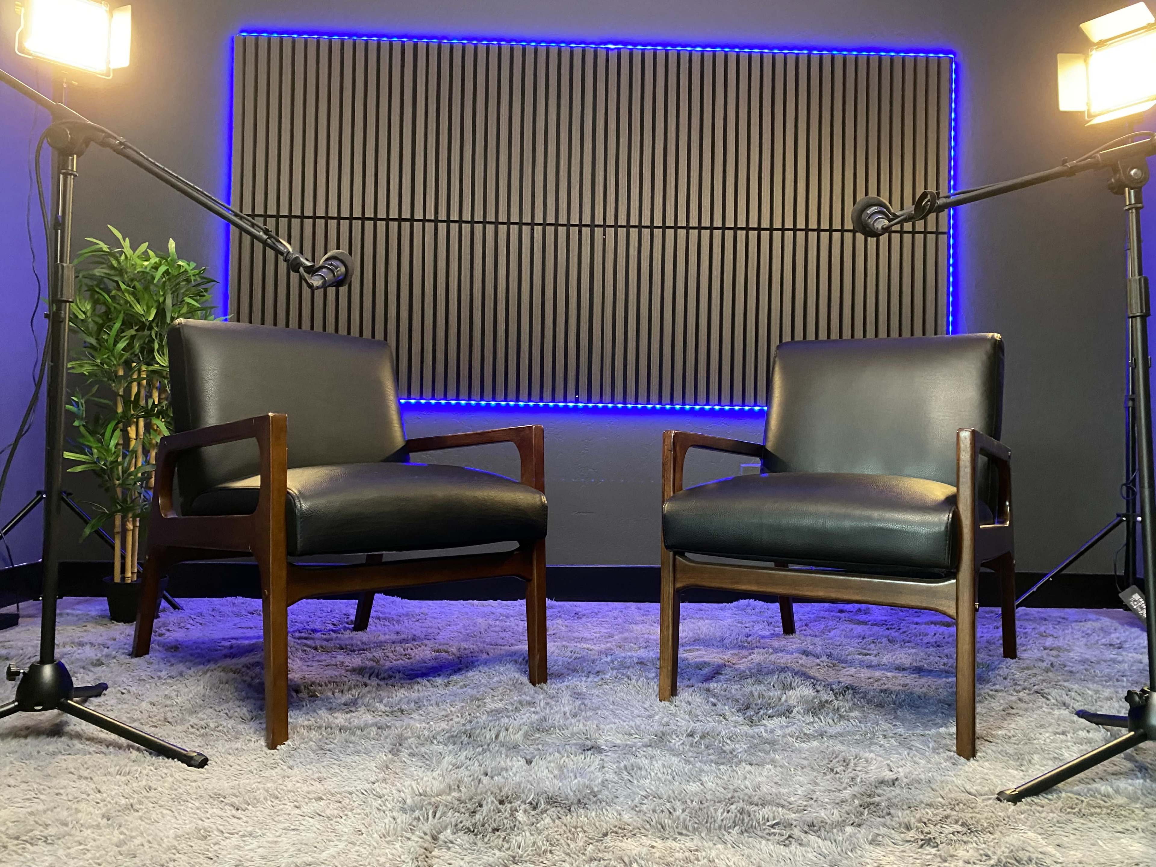 Two wooden chairs with black upholstery positioned in front of a soundproof wall, illuminated by two studio lights and flanked by microphones.