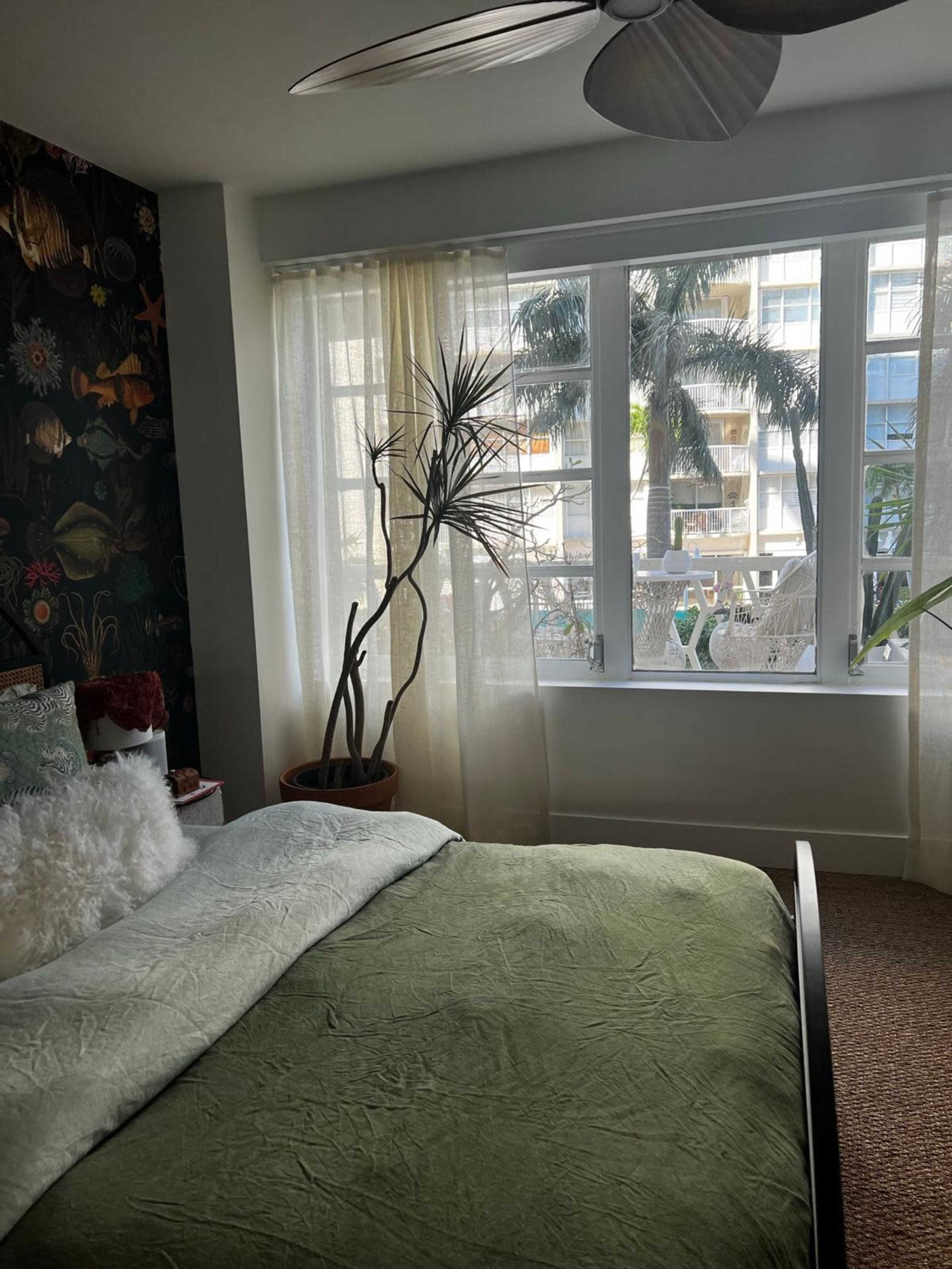 A bedroom featuring a green bedspread, a large window with flowing curtains, and a potted plant beside the bed.
