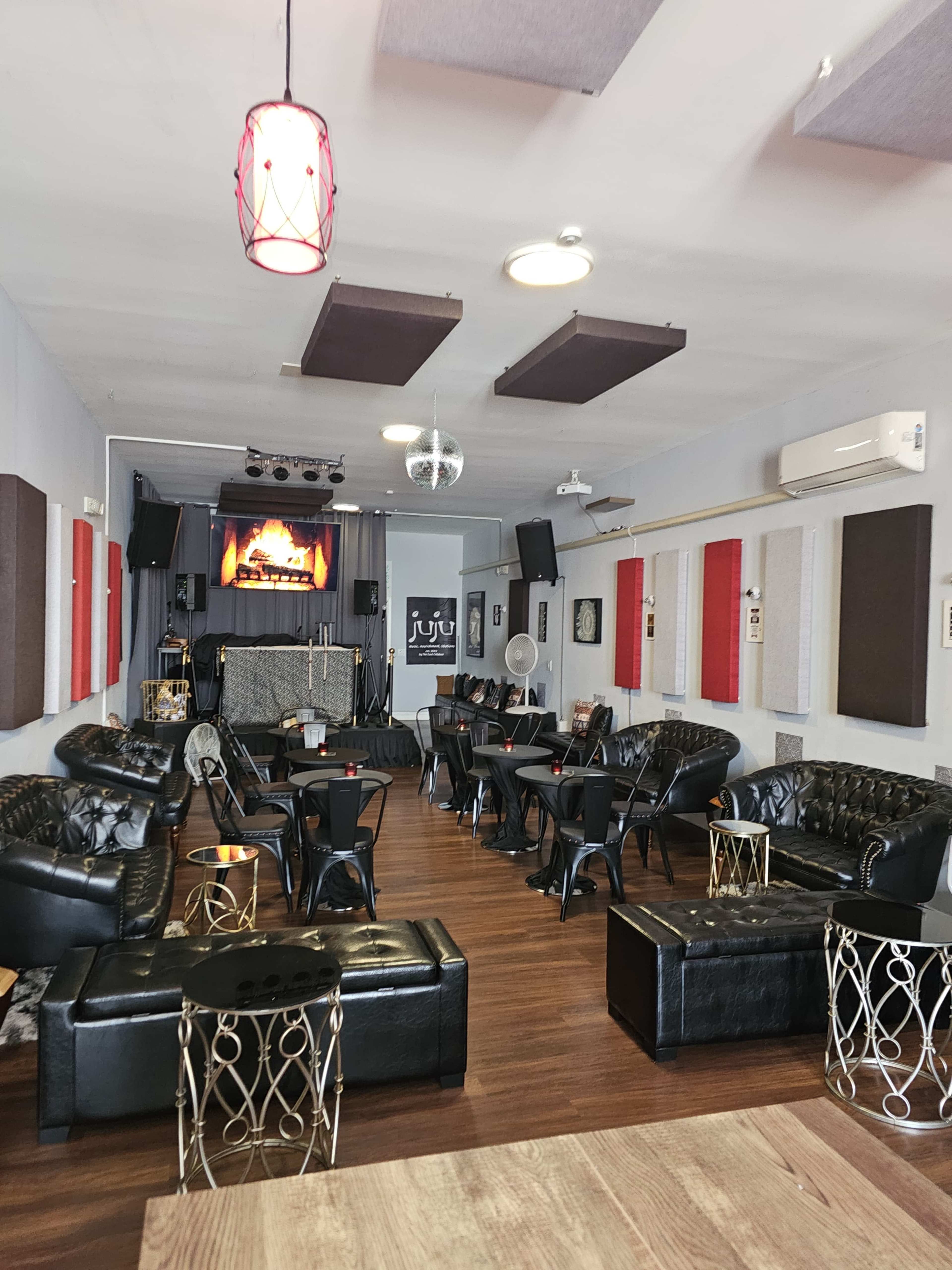 A modern lounge with black leather seating arranged around small tables and a stage at the front featuring a large screen.
