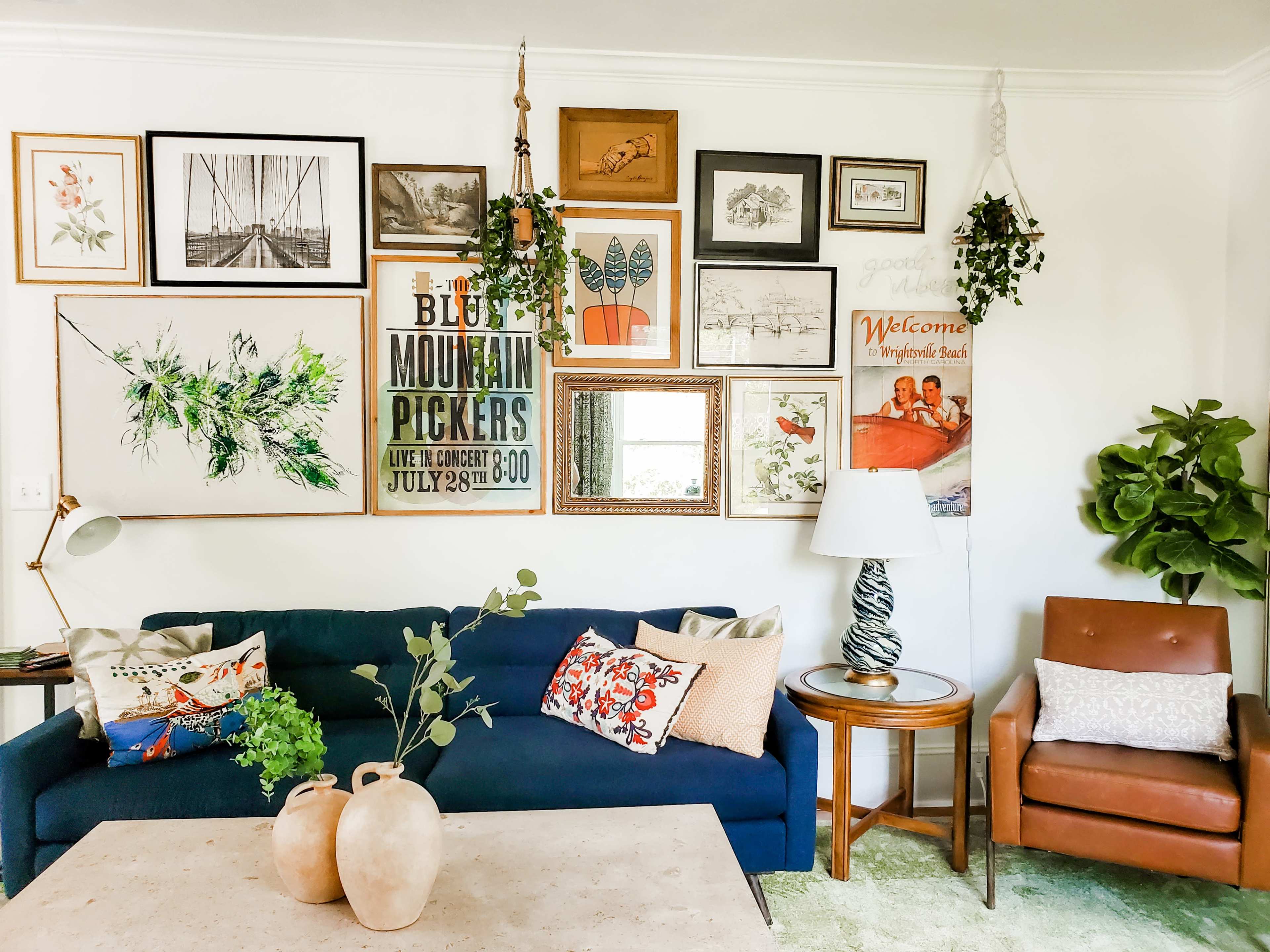A living room with a blue sofa adorned with decorative pillows, a wooden side table beside a brown leather armchair, and a gallery wall filled with various framed artworks and plants.