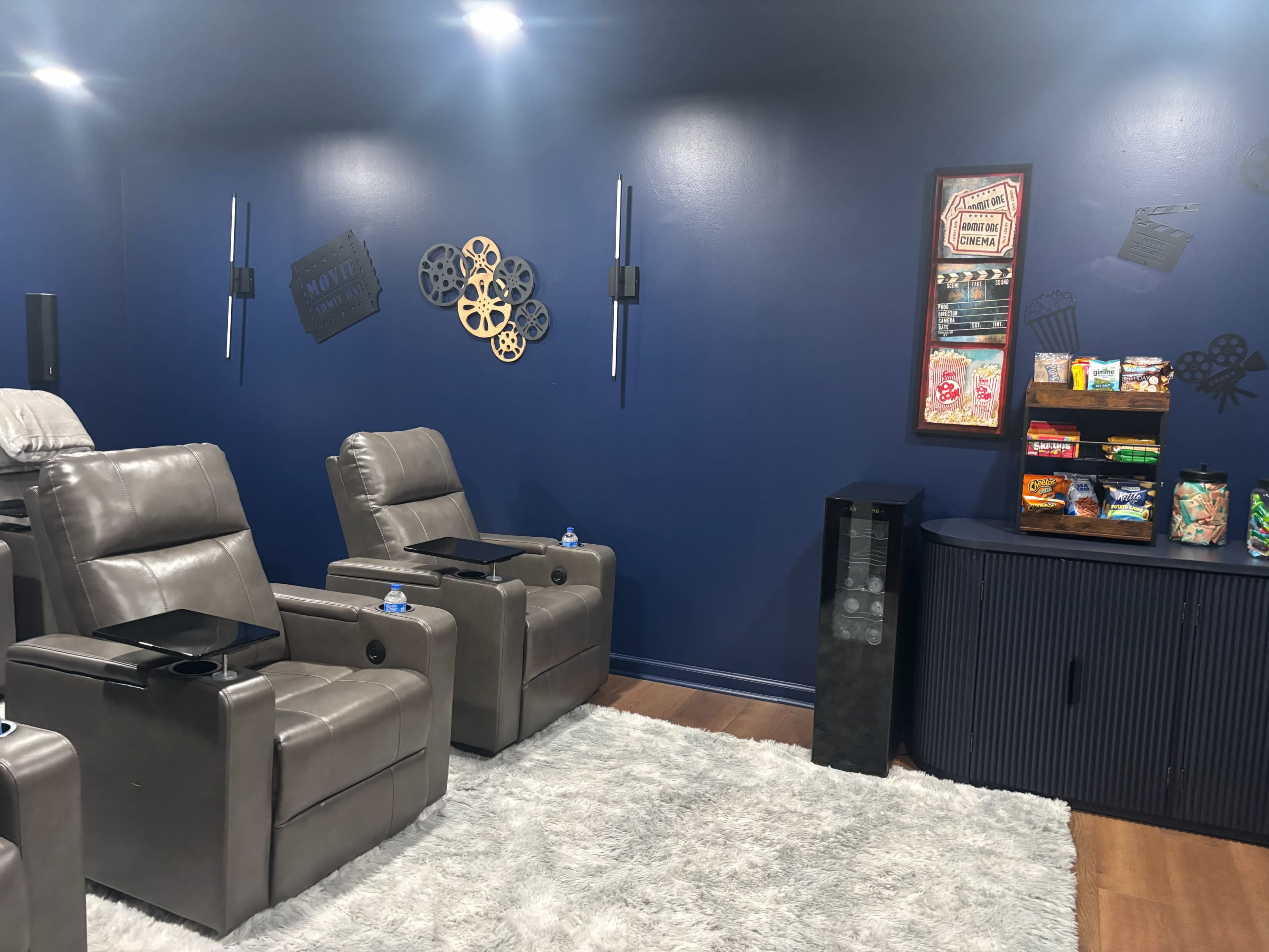 The image shows a cozy home theater with leather recliners, a dark blue wall, and a snack station featuring various treats.
