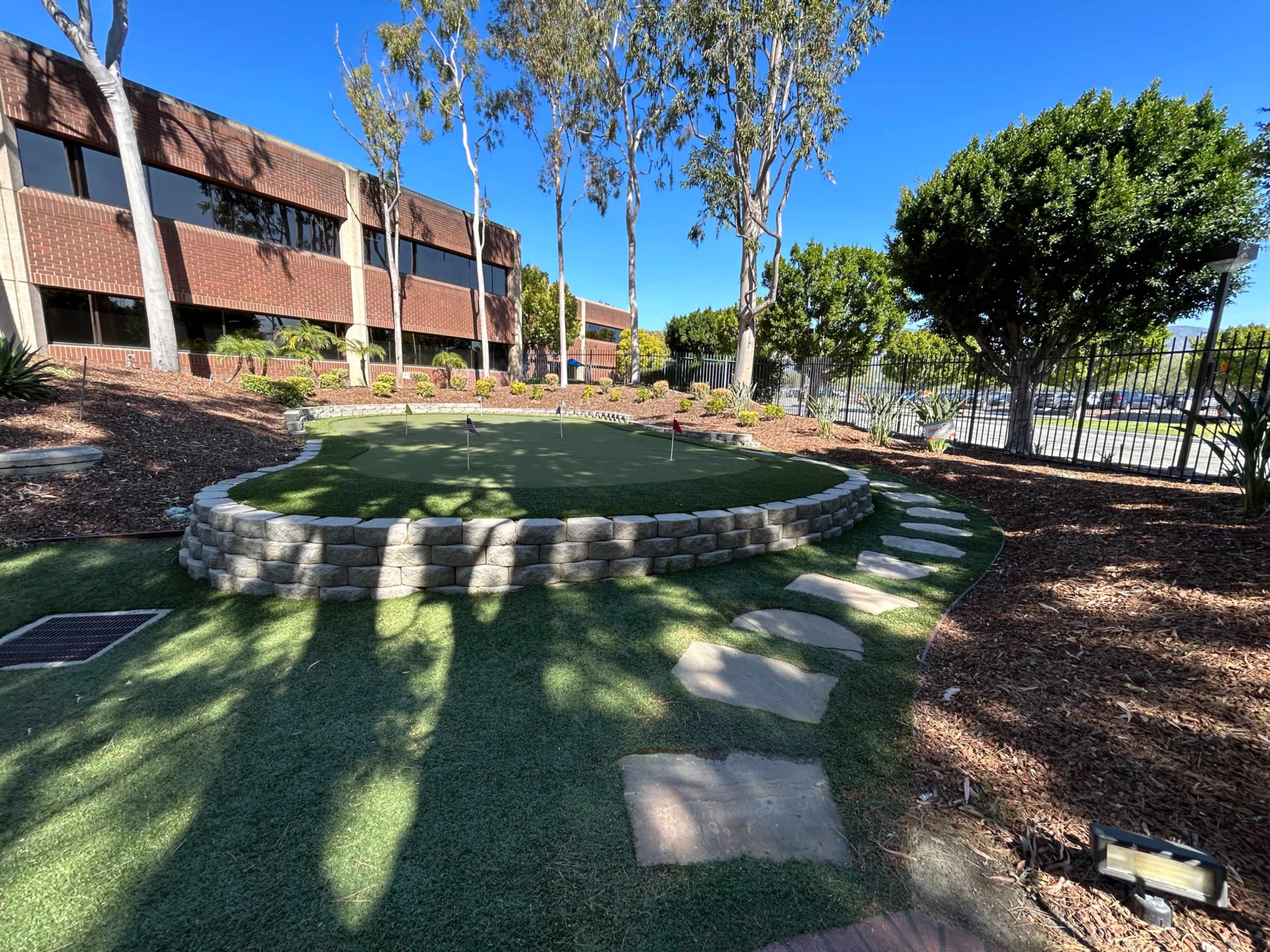 A landscaped area with a circular putting green surrounded by stone edging, palm trees, and a pathway of stone pavers.