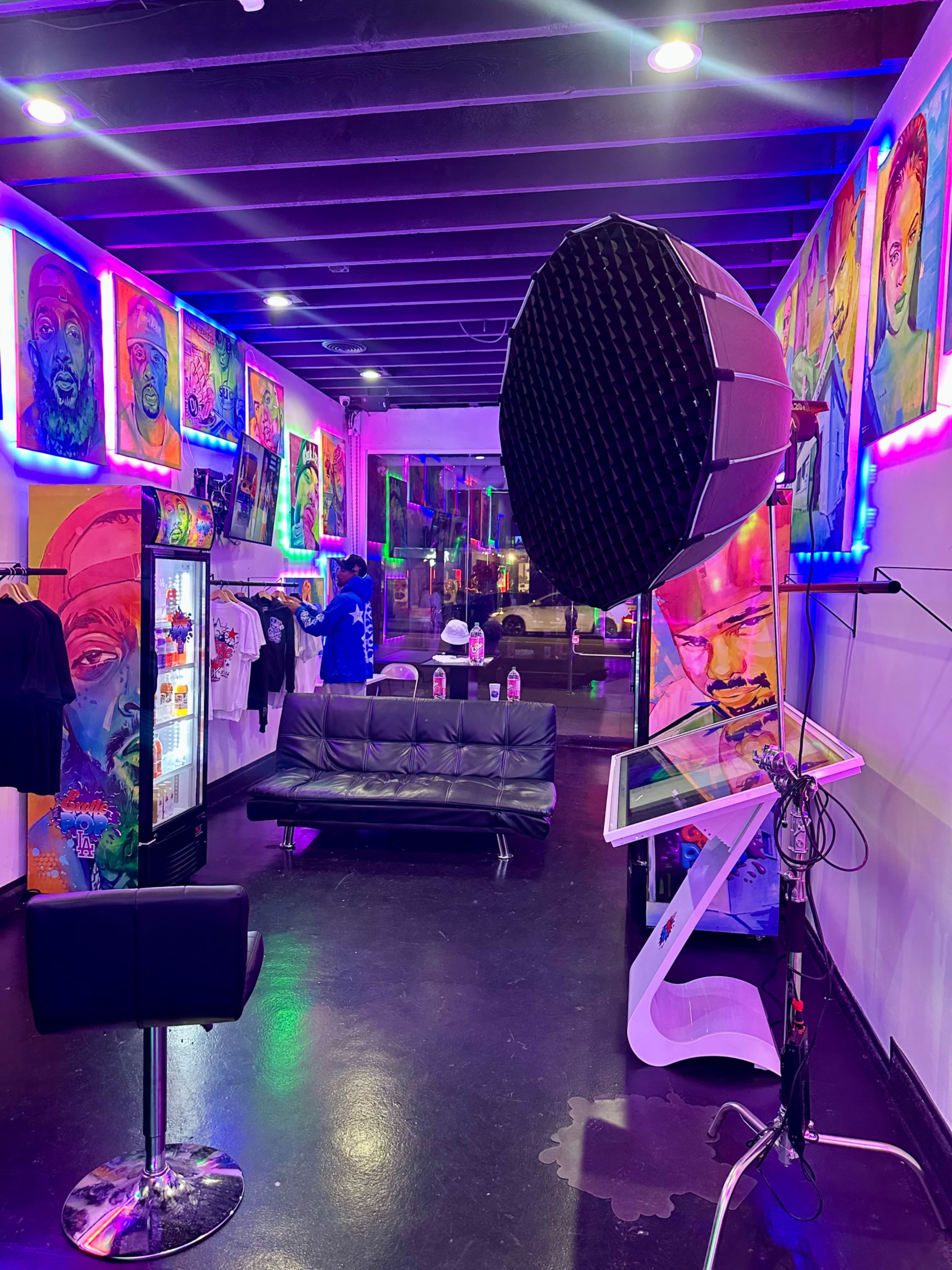 The image shows a colorful art studio featuring vibrant portraits on the walls, a black couch in the center, and a display of merchandise including T-shirts and a beverage fridge.