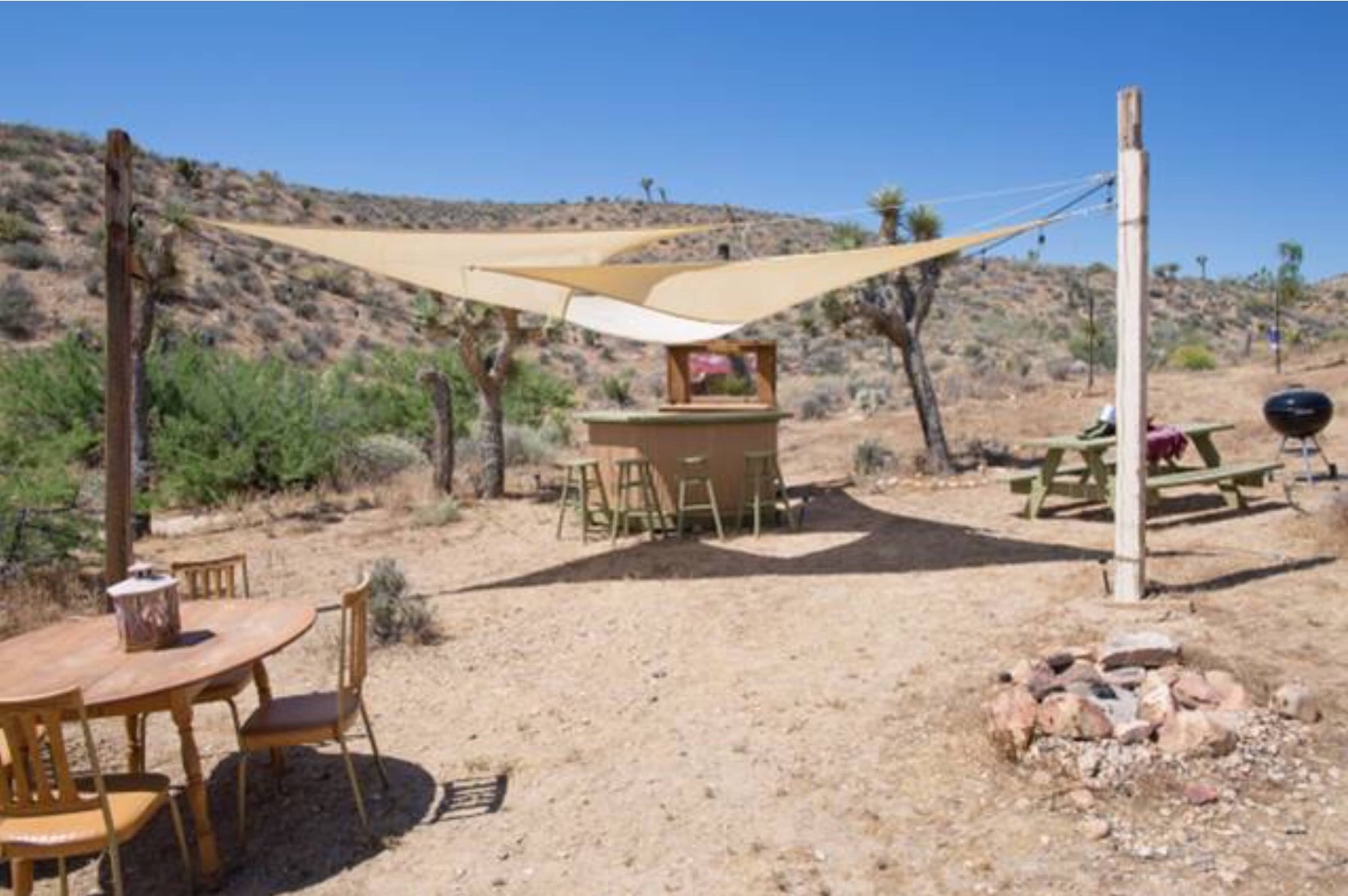 A shaded outdoor area features a wooden table and chairs, a bar, a picnic table, and a fire pit set against a desert landscape with sparse vegetation and distant hills.
