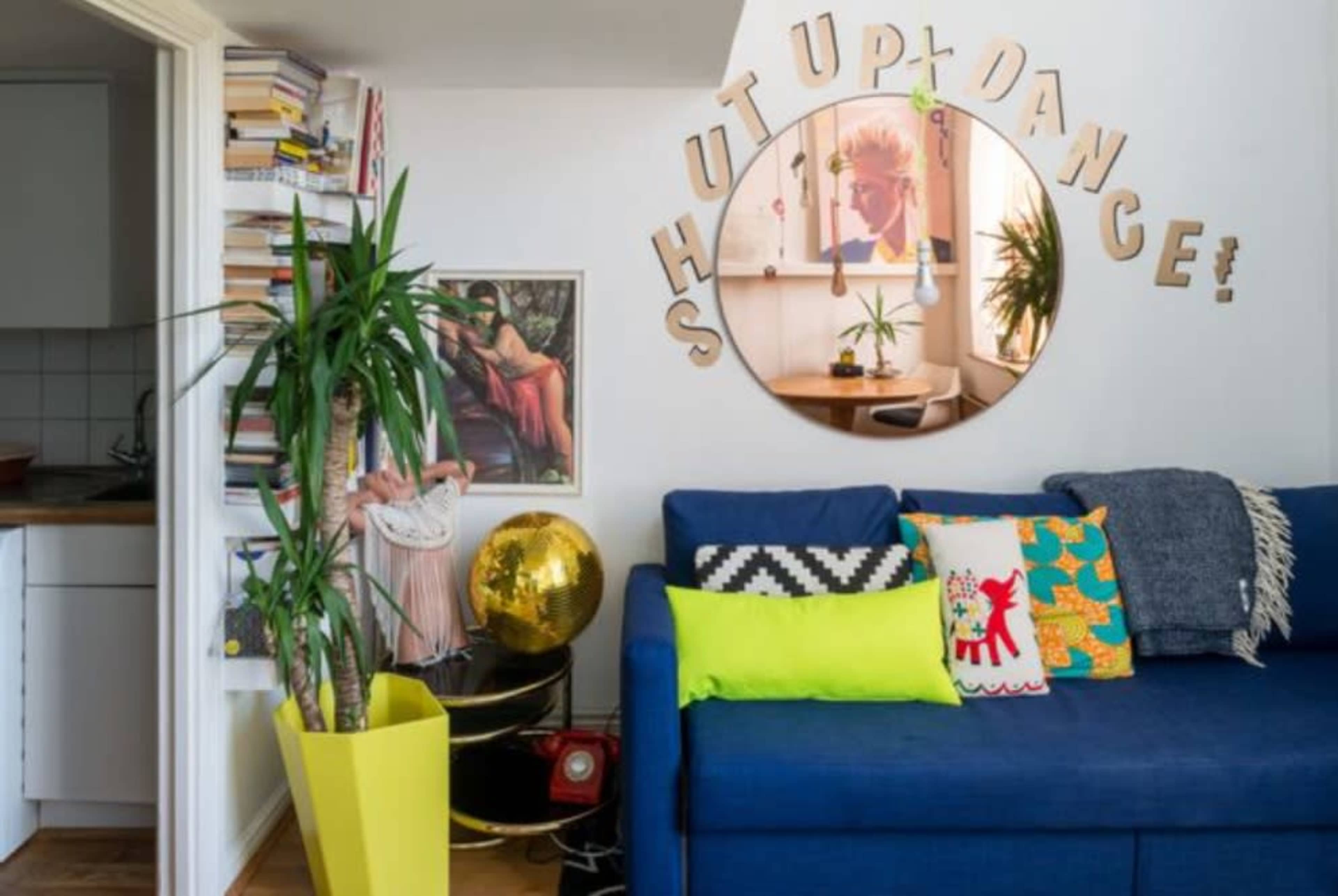 The image shows a corner of a living room featuring a blue sofa adorned with colorful pillows, a large round mirror with decorative lettering, and a yellow planter with a tall plant.