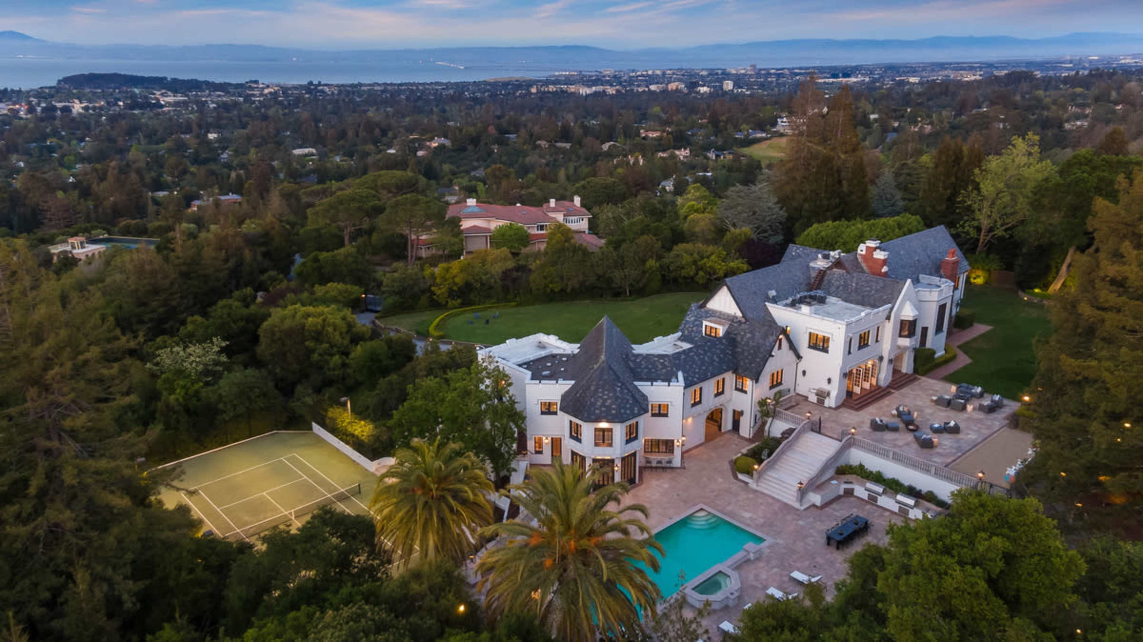 The image shows a large mansion surrounded by trees, with a swimming pool and a tennis court in the backyard.