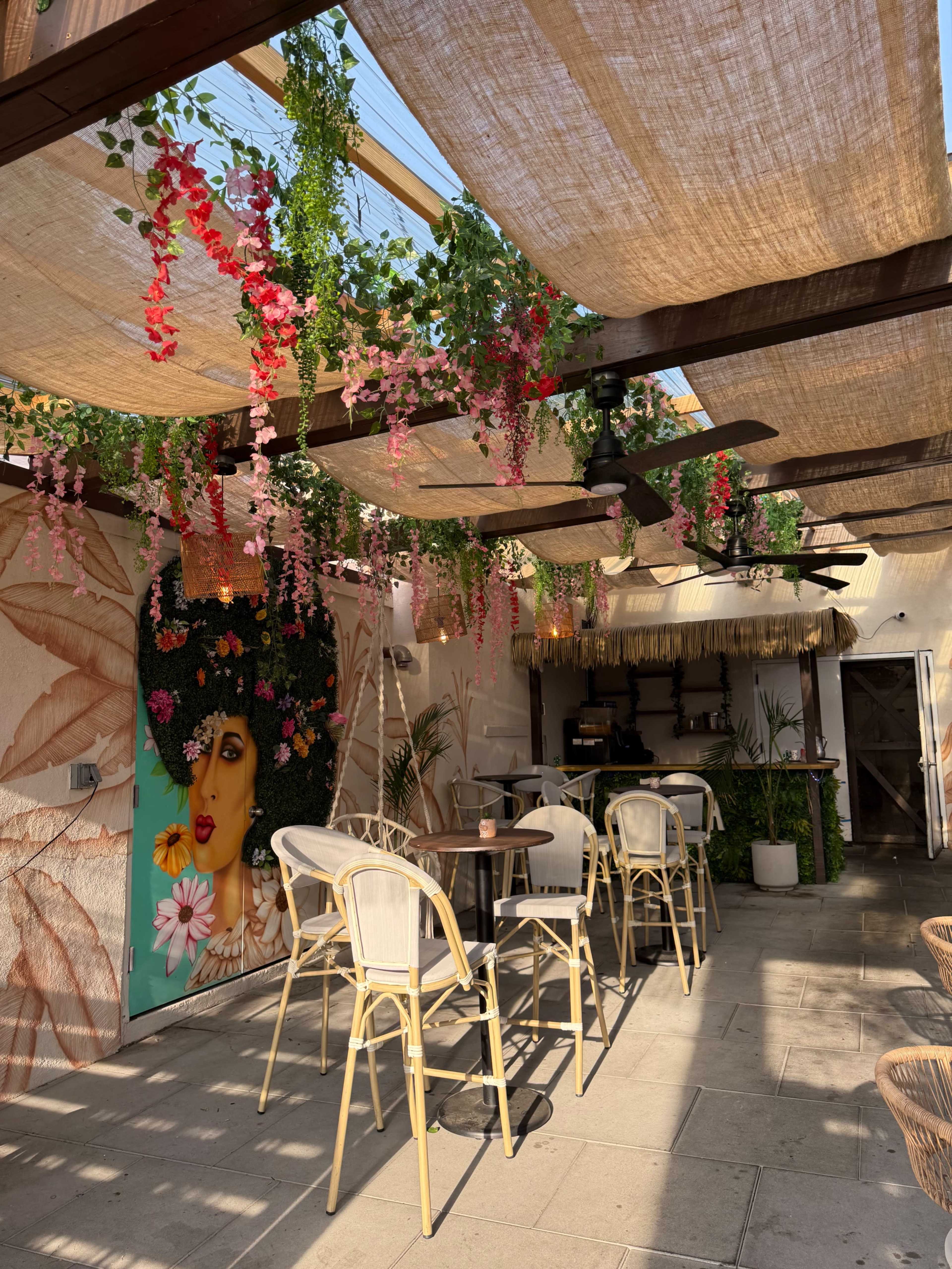 The image shows a vibrant outdoor café space adorned with hanging flowers, art on the wall, and seating arranged under large fabric canopies.