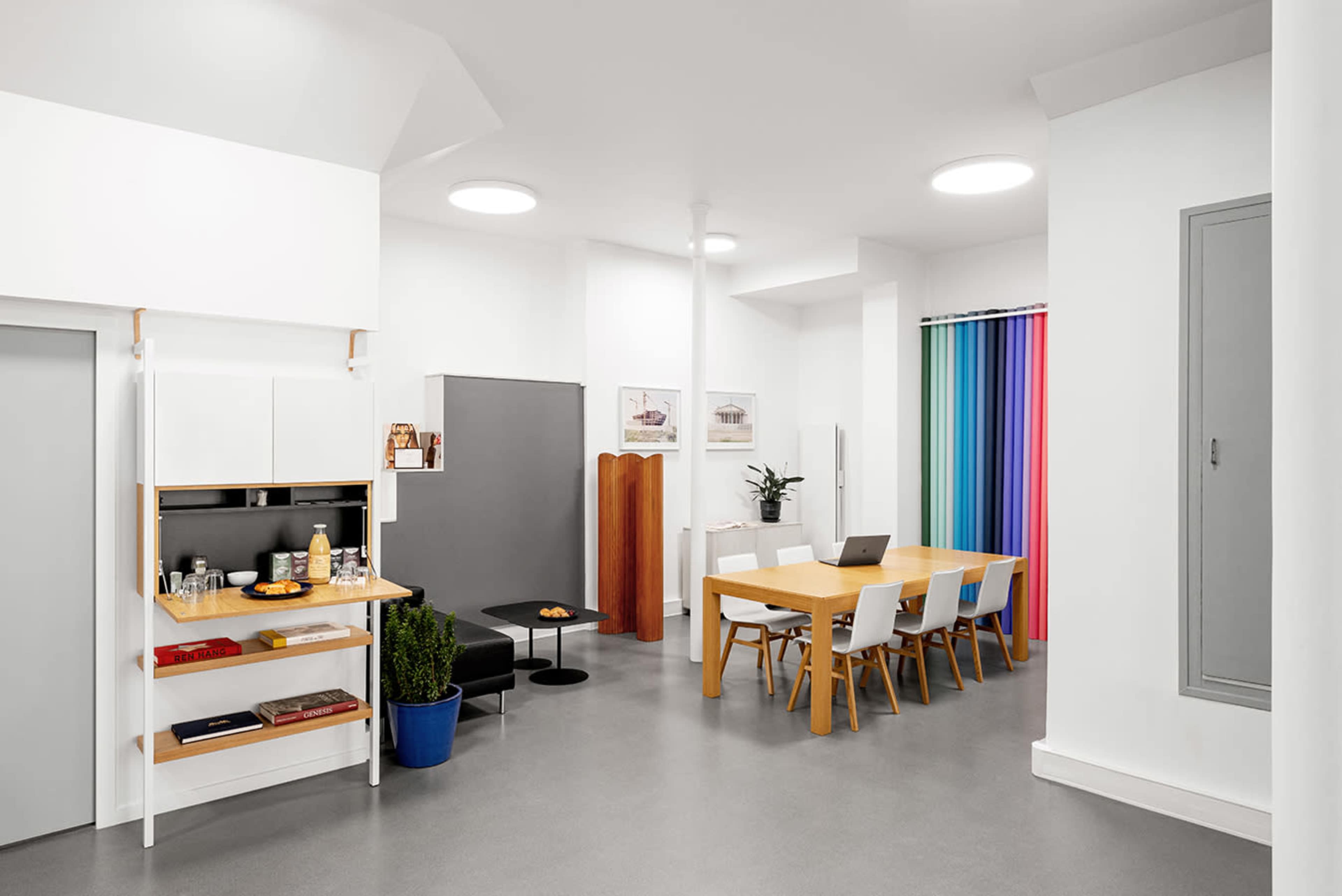 A modern, minimalist kitchen and dining area with a wooden table, white chairs, and a colorful curtain in the background.