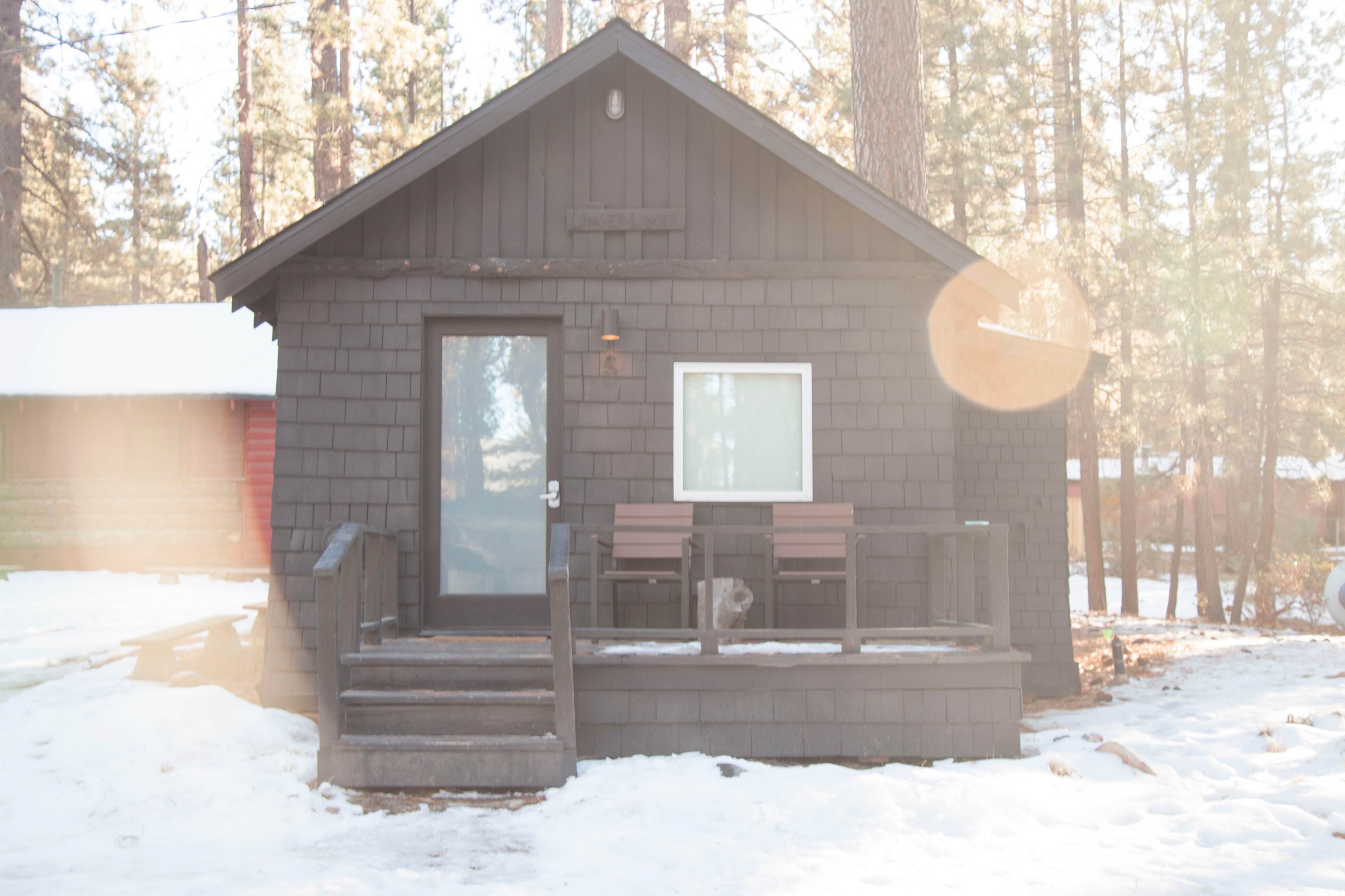A small, dark-colored cabin with a front porch and steps is surrounded by snow and tall trees.