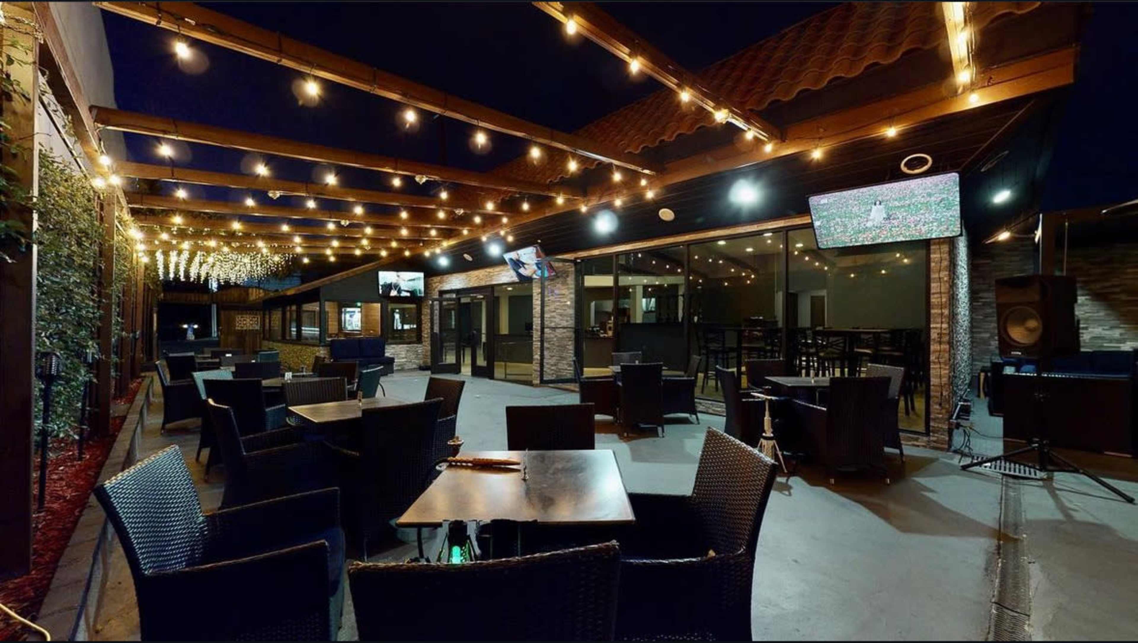 The image shows an outdoor dining area of a restaurant, featuring dark wicker tables and chairs, string lights overhead, and several flat-screen TVs mounted on the walls.