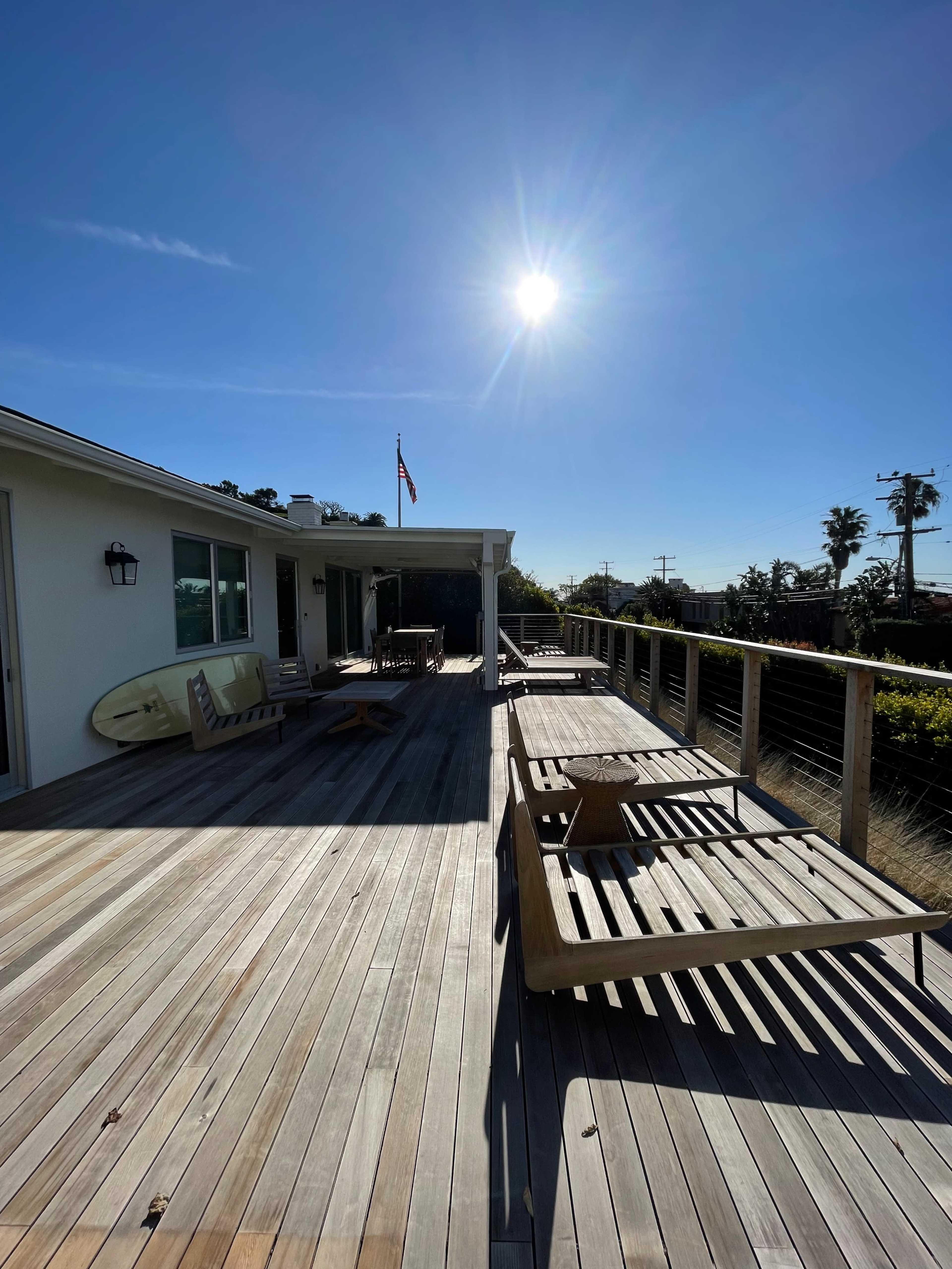 The image shows a wooden deck with tables and chairs, a surfboard leaning against the house, and a clear blue sky with the sun shining brightly overhead.
