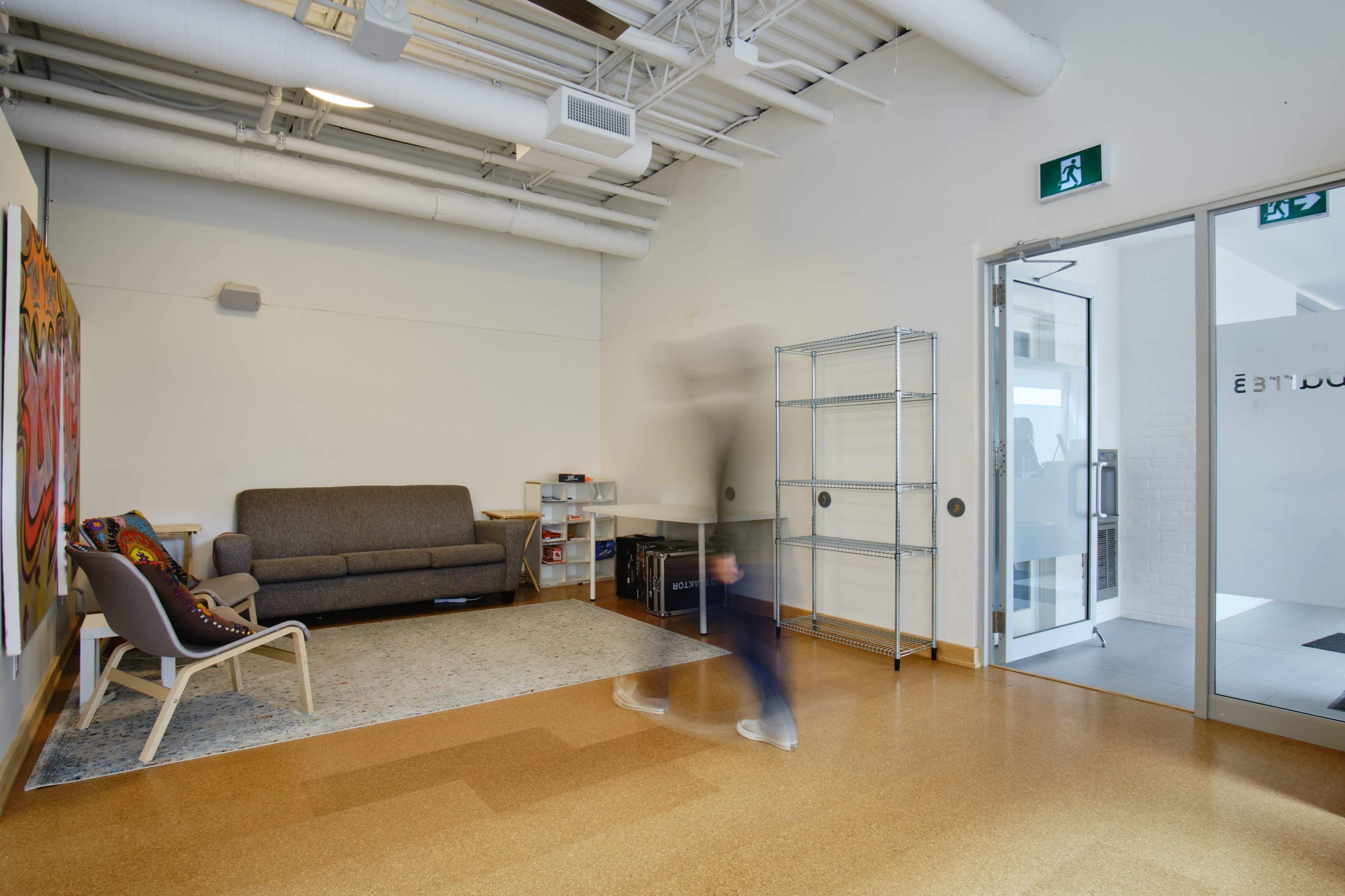 A person walks through a minimalist waiting area with a couch, shelving unit, and an exit door.