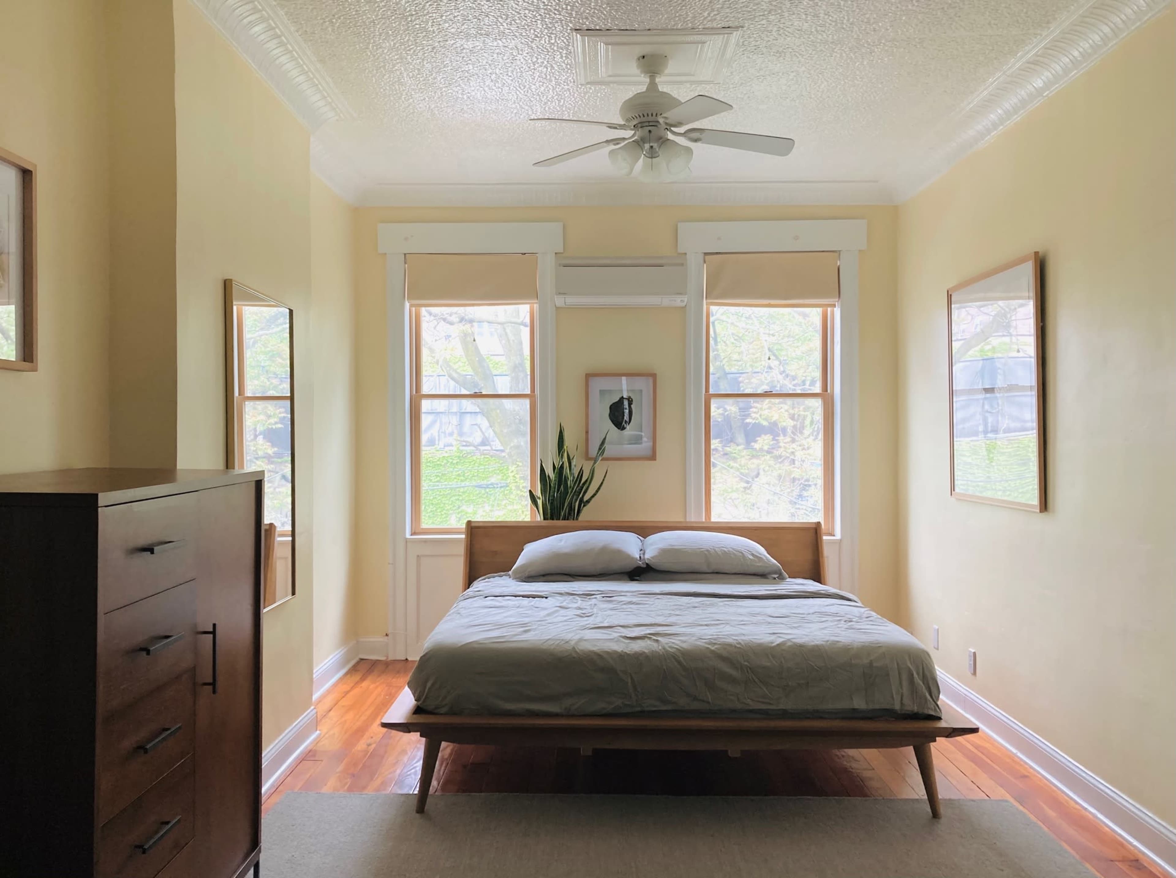 The image shows a bright bedroom featuring a wooden bed with white bedding, a large dresser, and two windows framed by light curtains.
