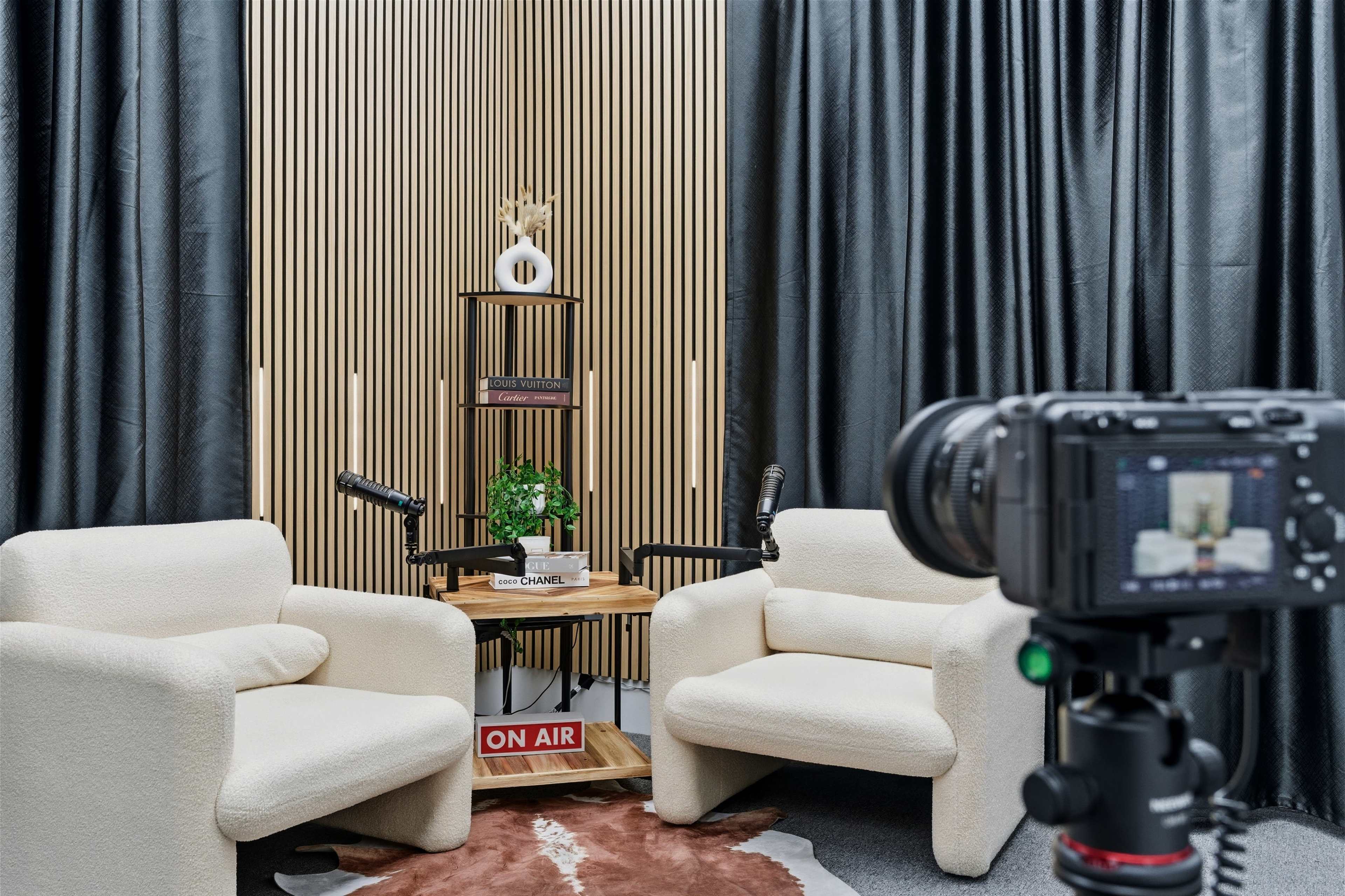 The image shows a podcasting setup with two white armchairs, a small wooden table, and a camera positioned to capture the scene, all set against a backdrop of black curtains and wooden panels.