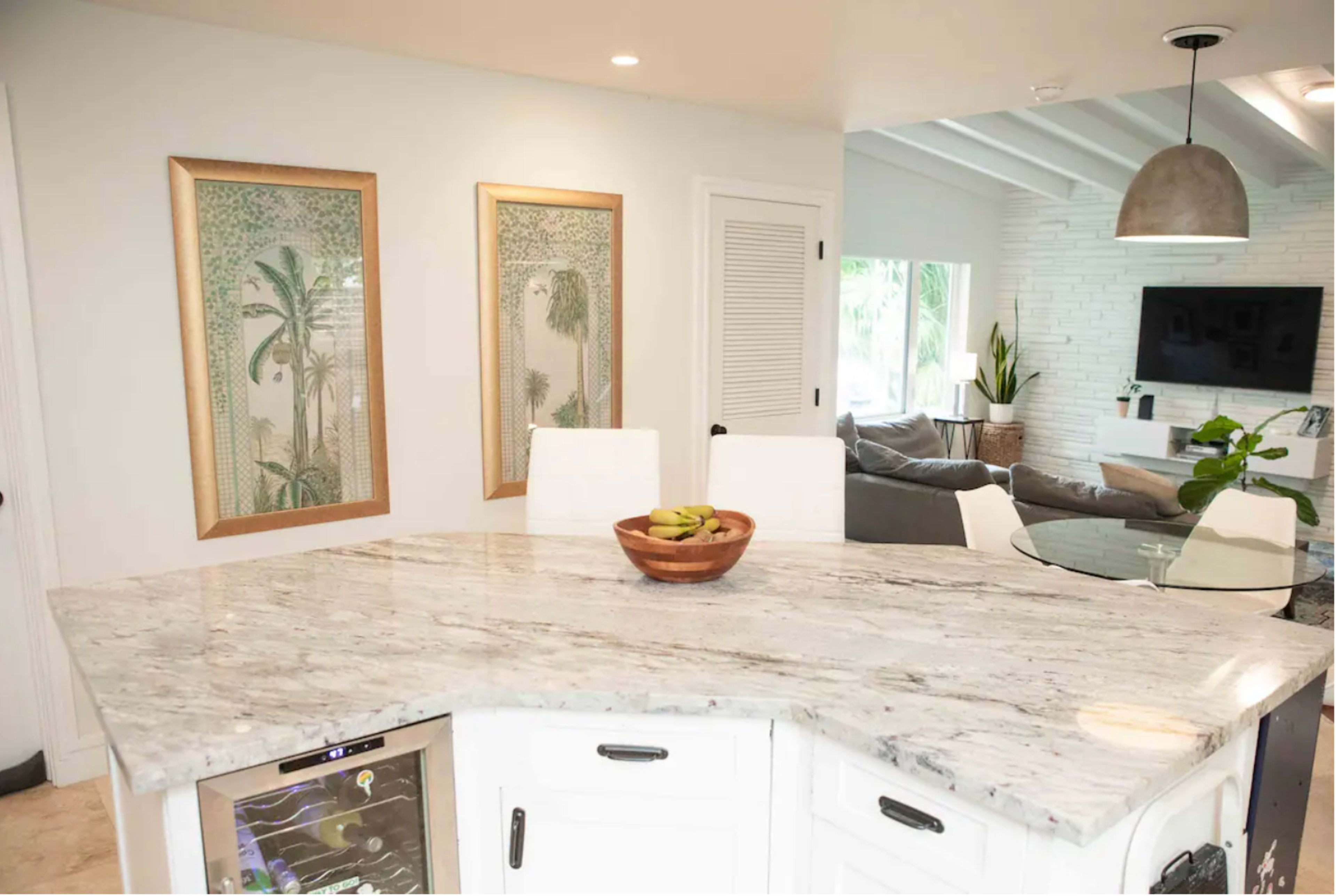 A kitchen countertop made of marble extends into an open living area, featuring two framed artworks on the wall and a bowl of fruit on the counter.