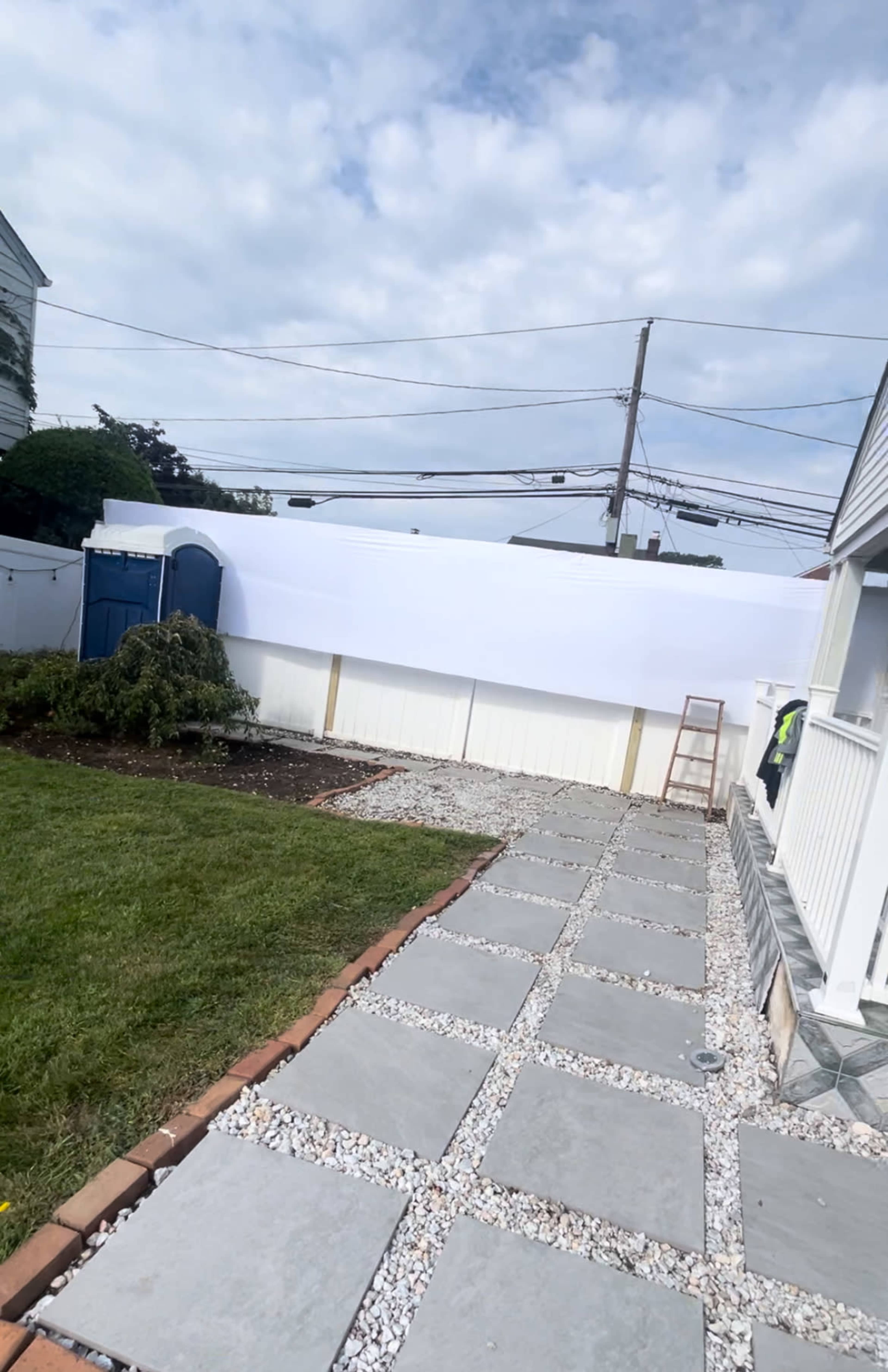 The image shows a backyard pathway made of gray stones leading to a white fence and a large white tarp or screen in the background, with some greenery and a ladder positioned nearby.