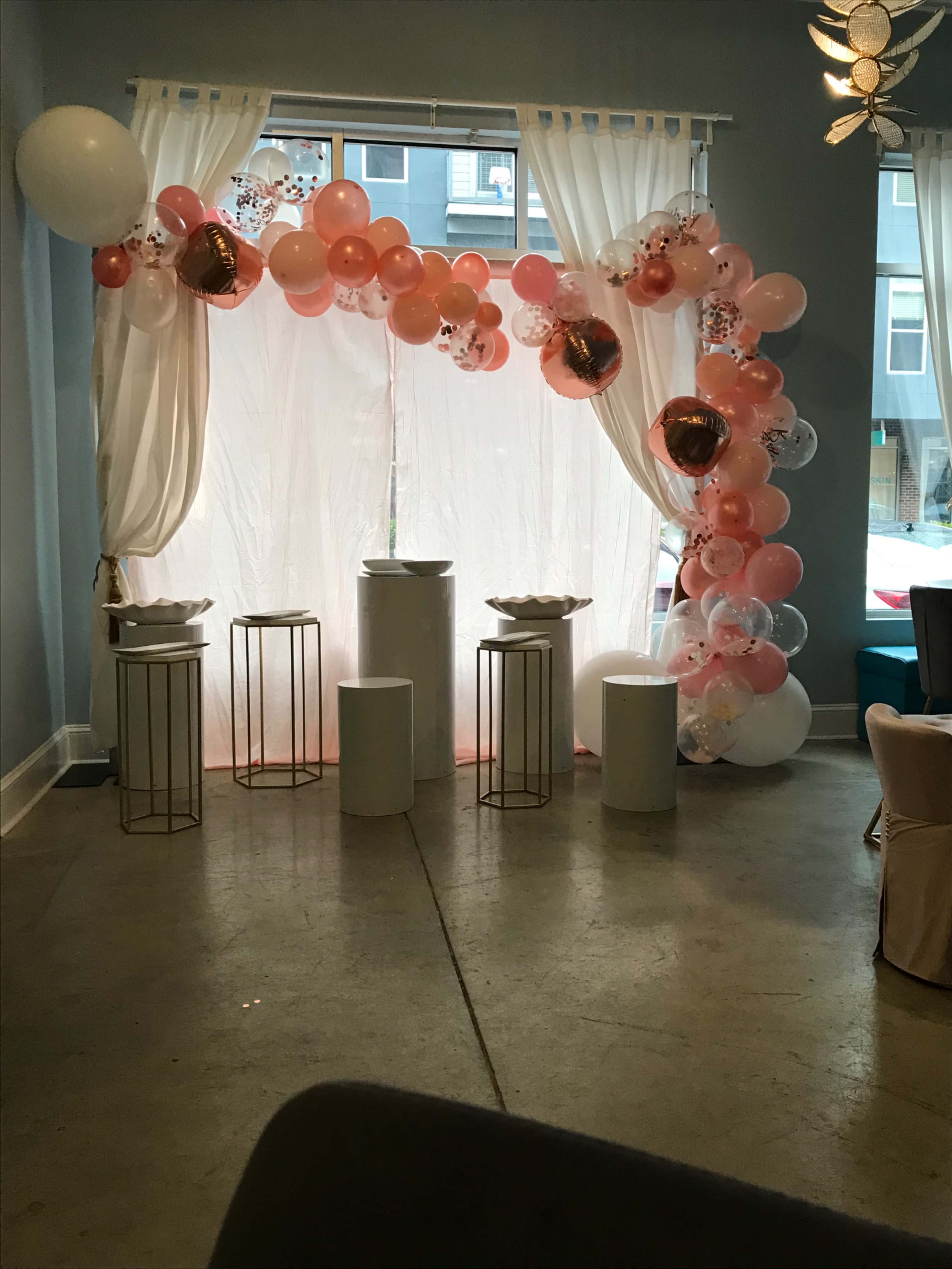 A decorated area with a balloon arch in shades of pink and white, draped curtains, and four white pedestals set up in front.
