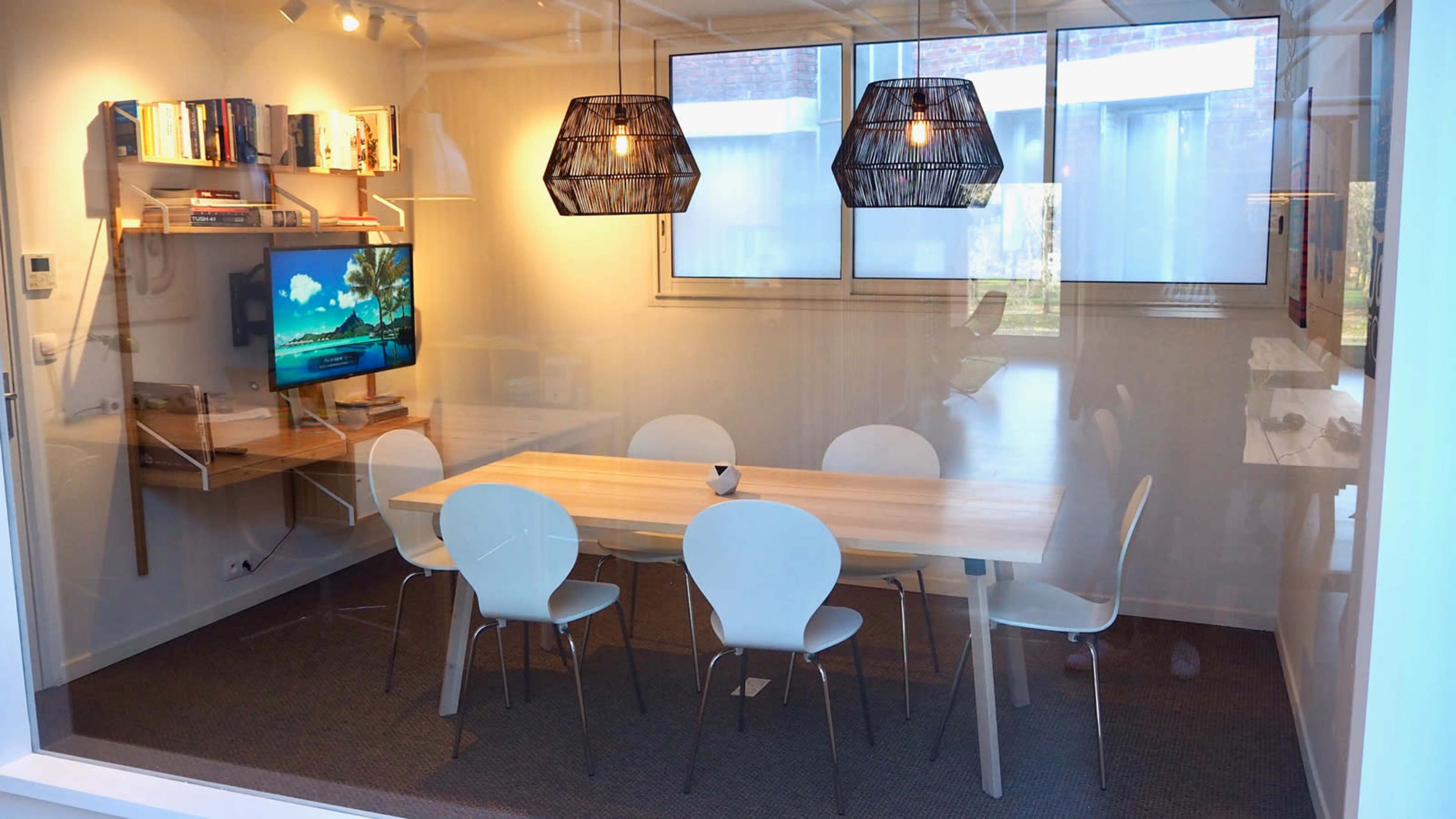 A modern conference room featuring a wooden table surrounded by white chairs, a wall-mounted television, and two pendant lights above the table.