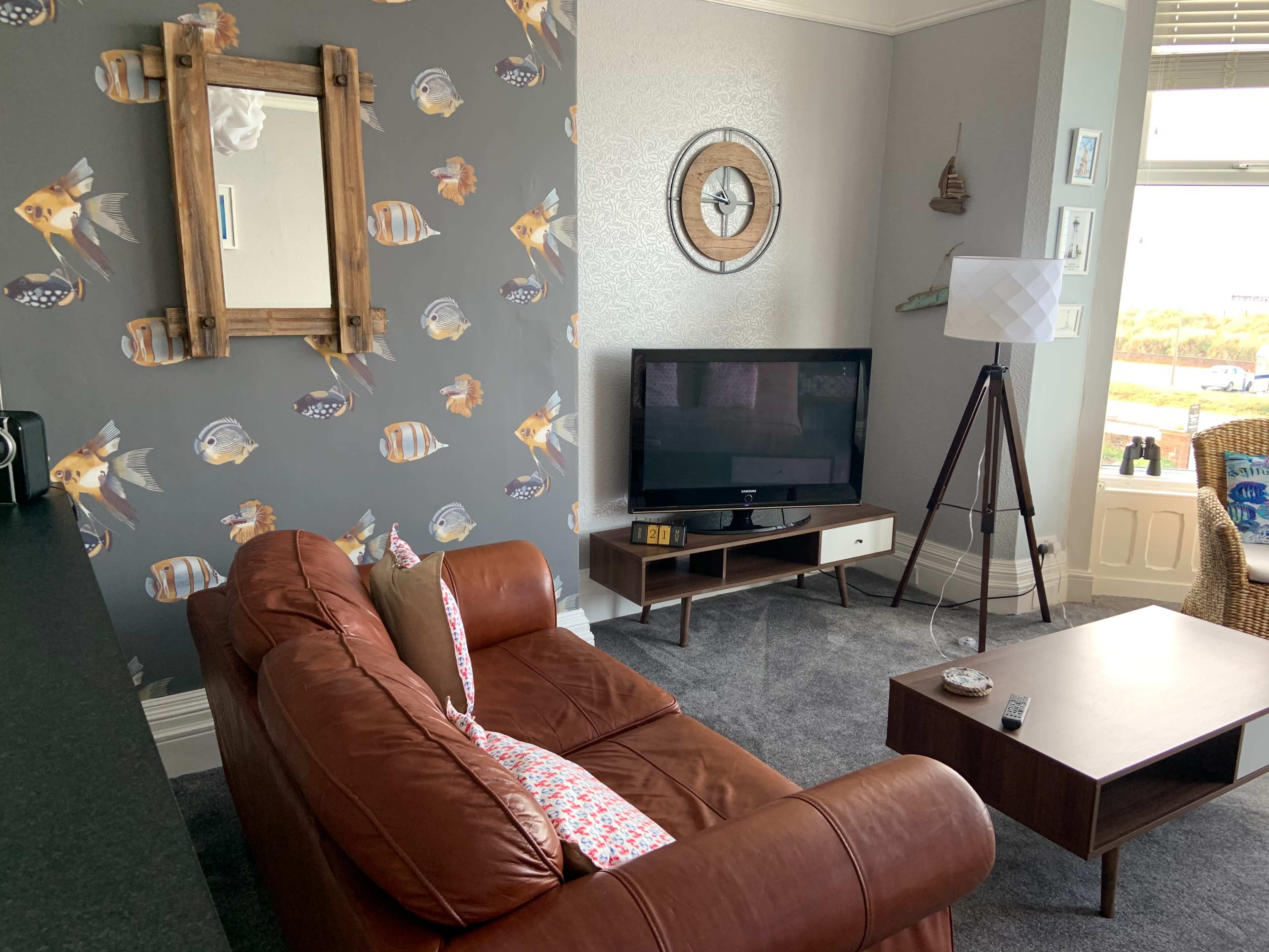 A living room with a brown leather couch, a wall with fish-patterned wallpaper, a television on a low cabinet, and a tall lamp beside a window.