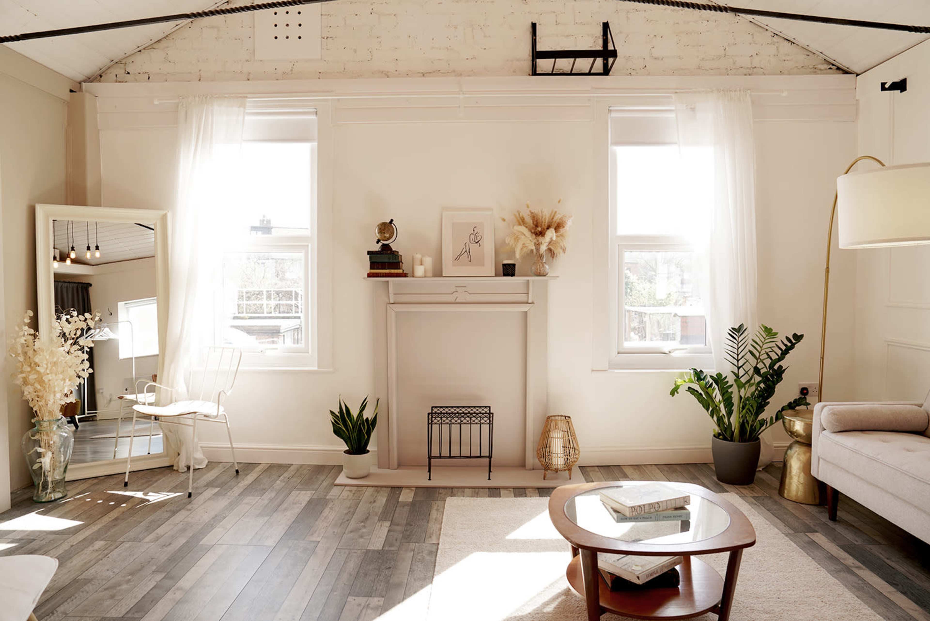 The image shows a bright, airy living room featuring large windows, a decorative fireplace, and a combination of plants and minimalist furniture.