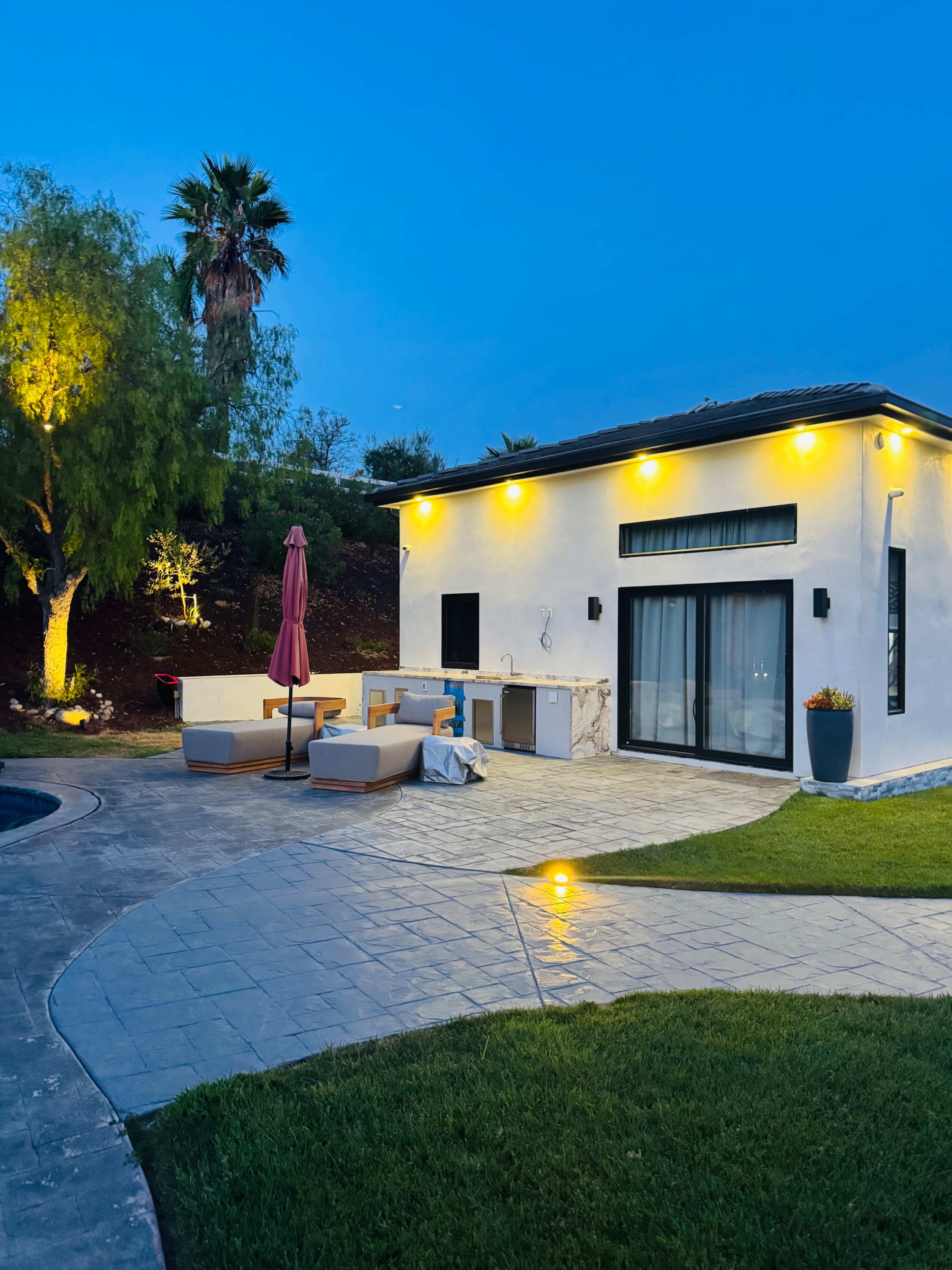 A well-lit outdoor patio features lounge furniture, an umbrella, and an outdoor kitchen beside a swimming pool, with palm trees in the background at dusk.